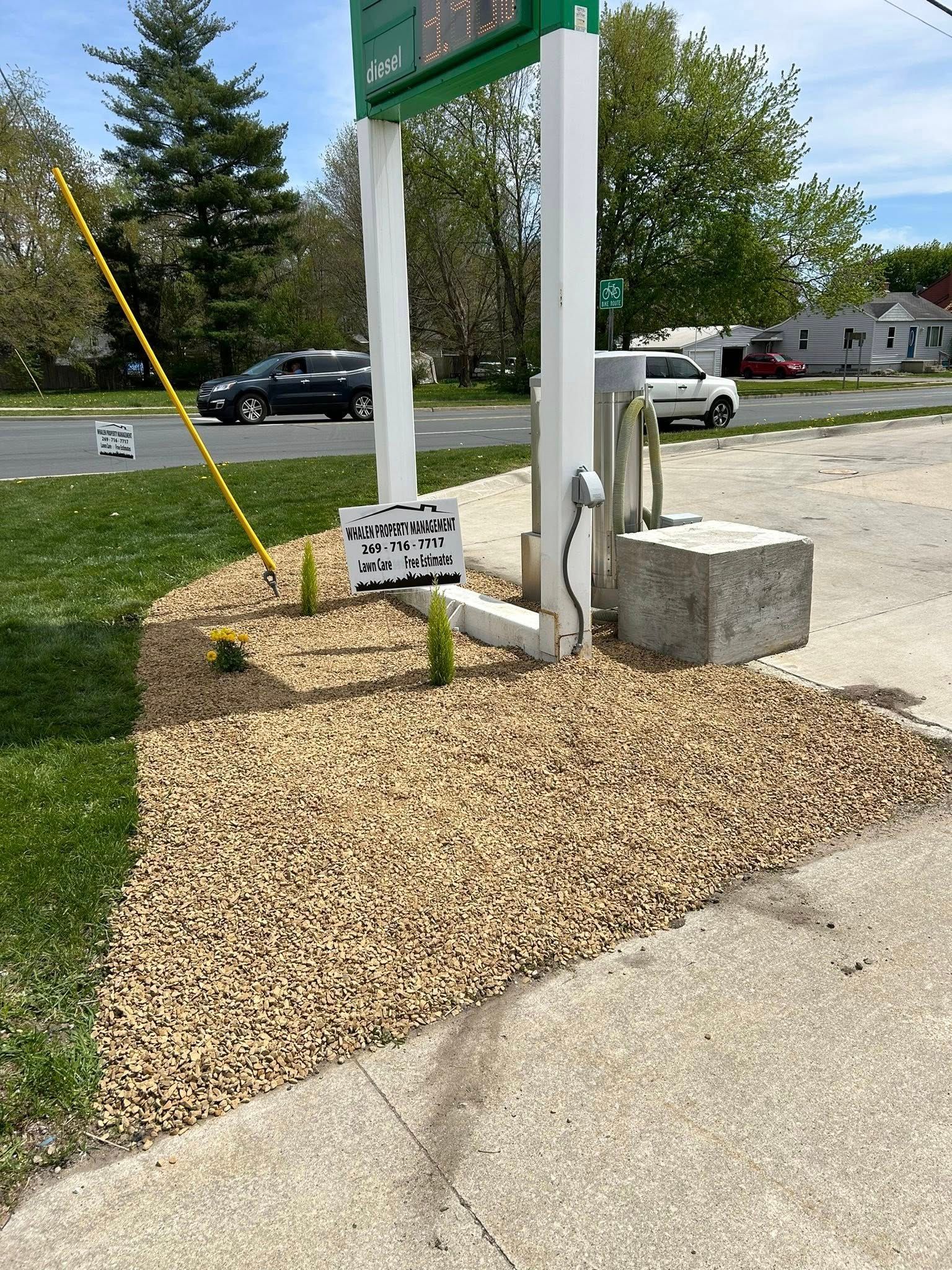 A small sign sits on a gravel patch near a gas station sign post and a concrete block.