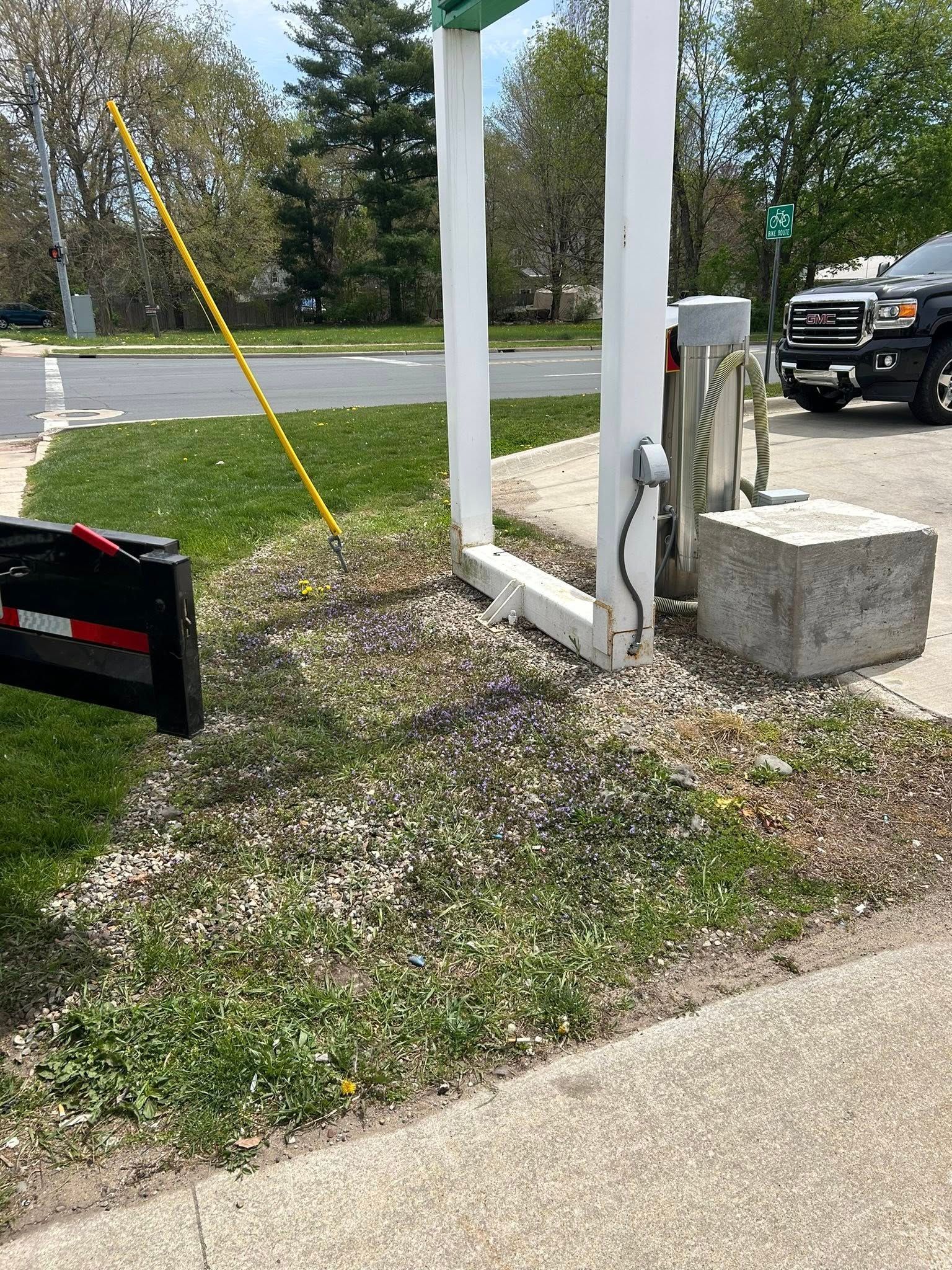 A yellow cable runs from a metal charging station across a gravel and grass patch near a parked truck and a concrete block.