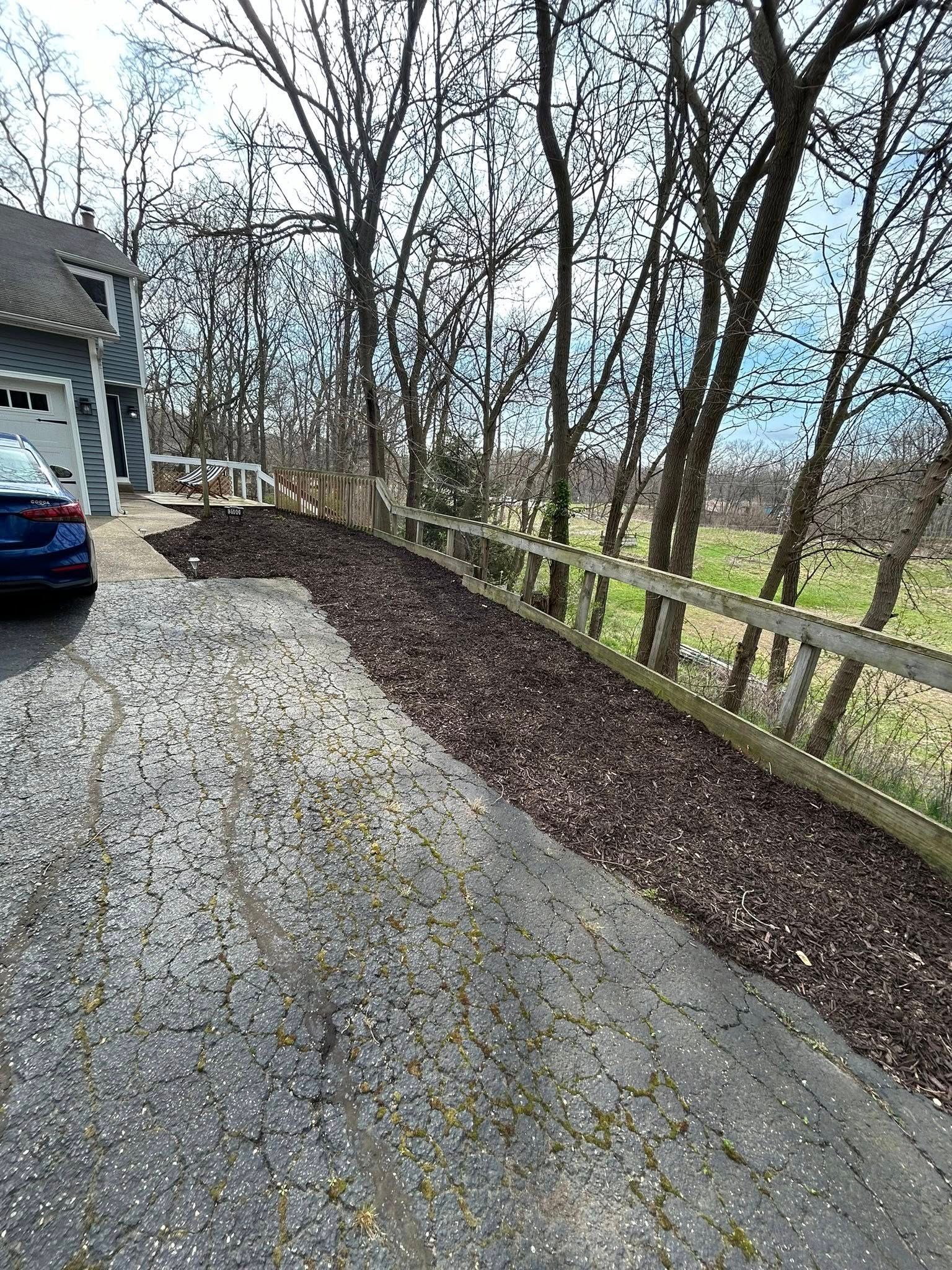 A gravel driveway bordered by a wooden fence, with a layer of fallen leaves and a blue car parked beside a house.