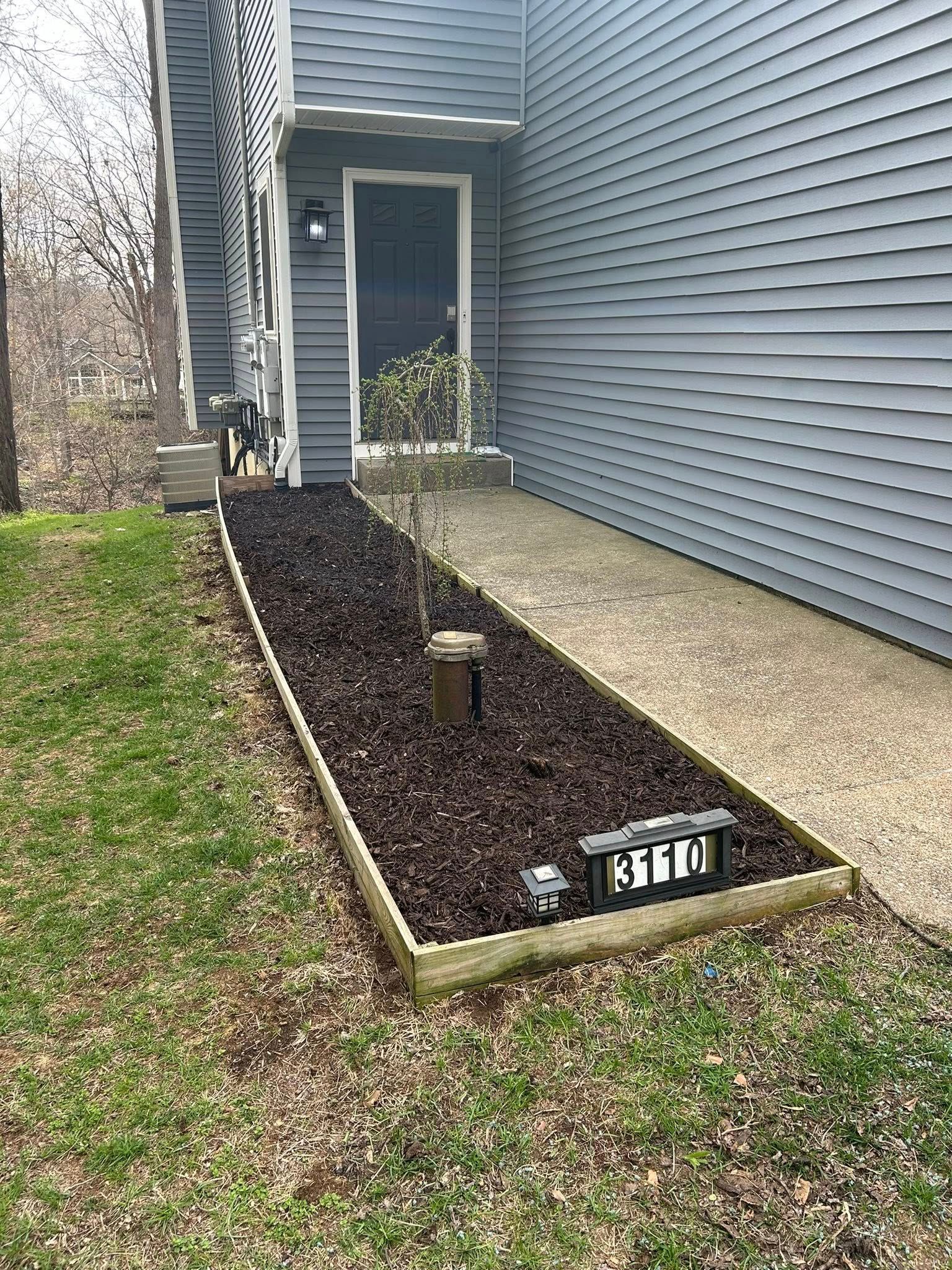 A gray siding house entrance features a rectangular garden bed with dark mulch, a small sapling, and a house number sign.