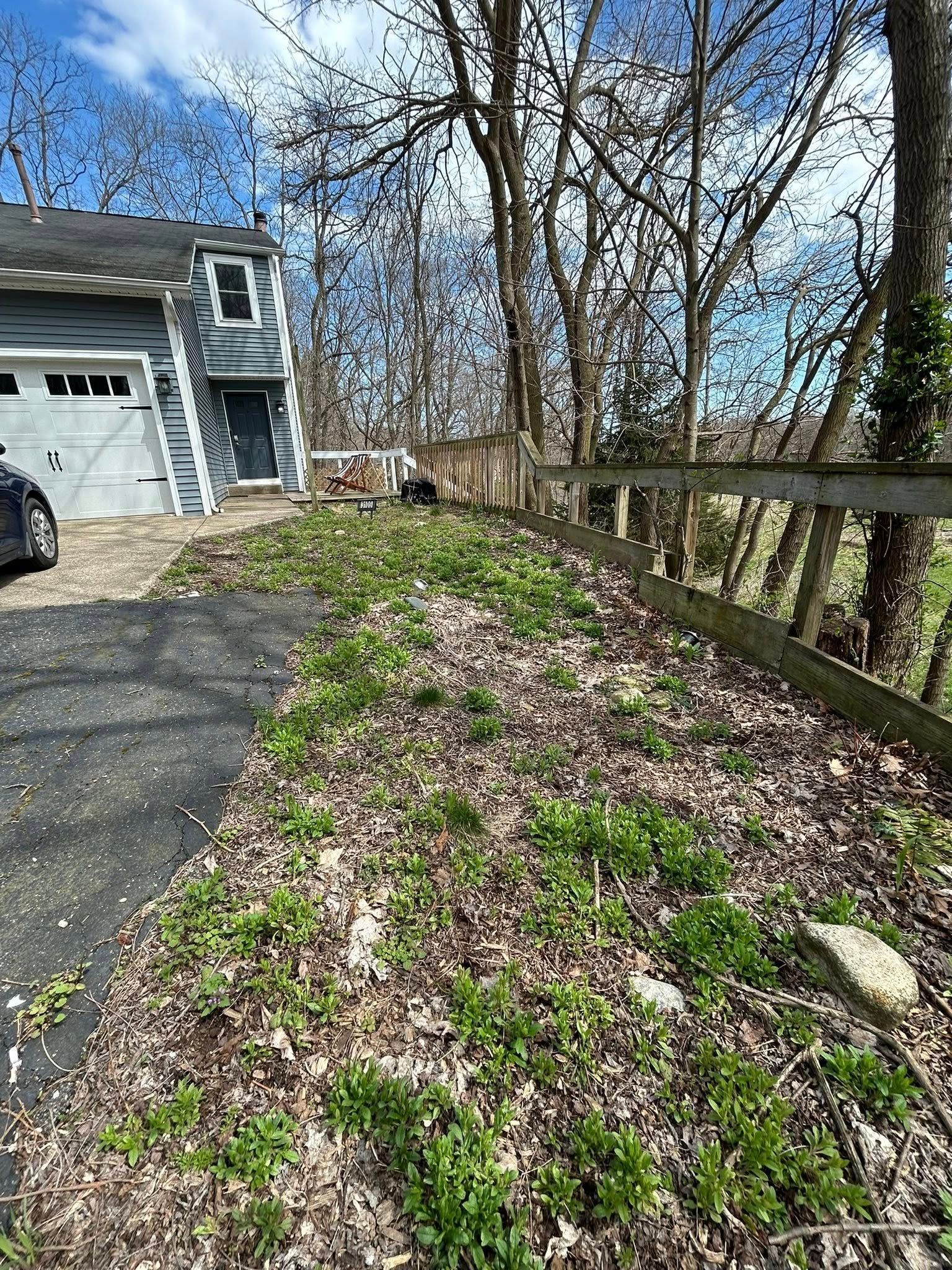 A grey house with a white garage sits next to a slope covered in dry leaves and small green plants along a wooden fence.