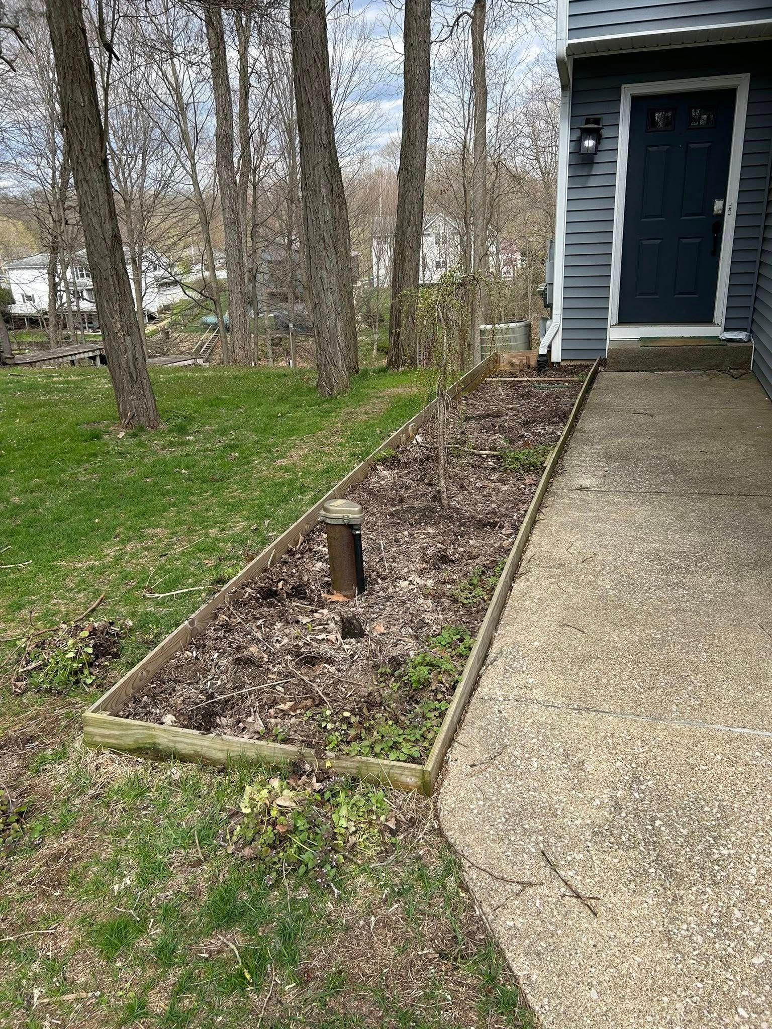 A rectangular garden bed with leaf mulch and a central post, bordering a concrete walkway beside a dark-sided house.