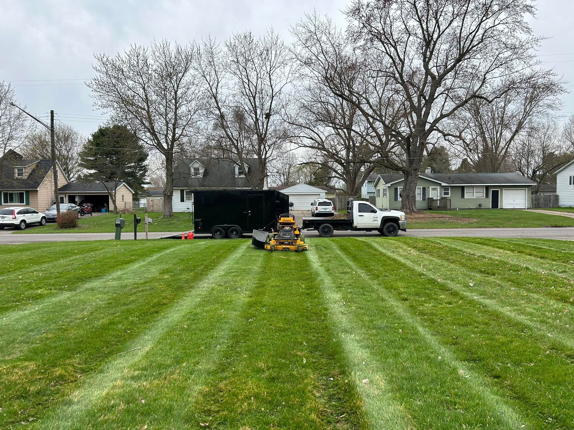 A yellow lawn mower creates stripes on a green lawn, with a black trailer and white truck parked on the street behind.