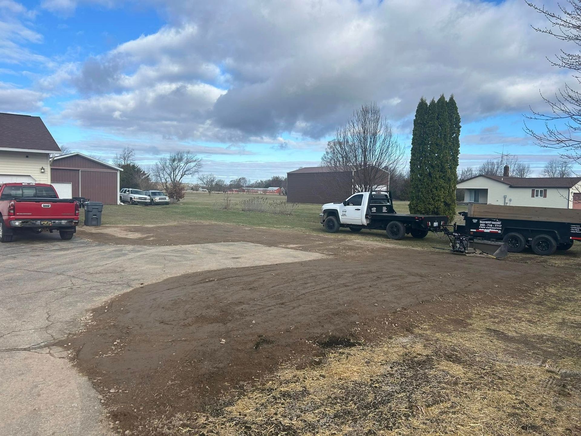 A gravel driveway with a red pickup truck, a white utility truck, and a trailer parked near houses on a cloudy day.