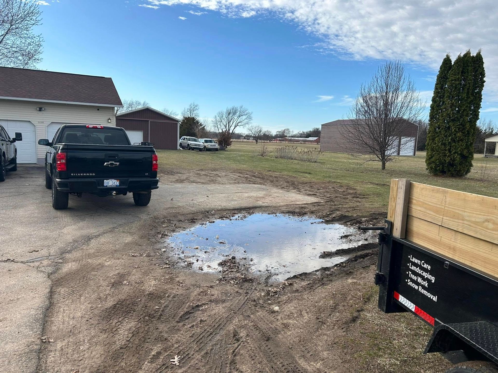A black pickup truck parked next to a large, muddy puddle in a gravel yard with buildings and trees in the background.