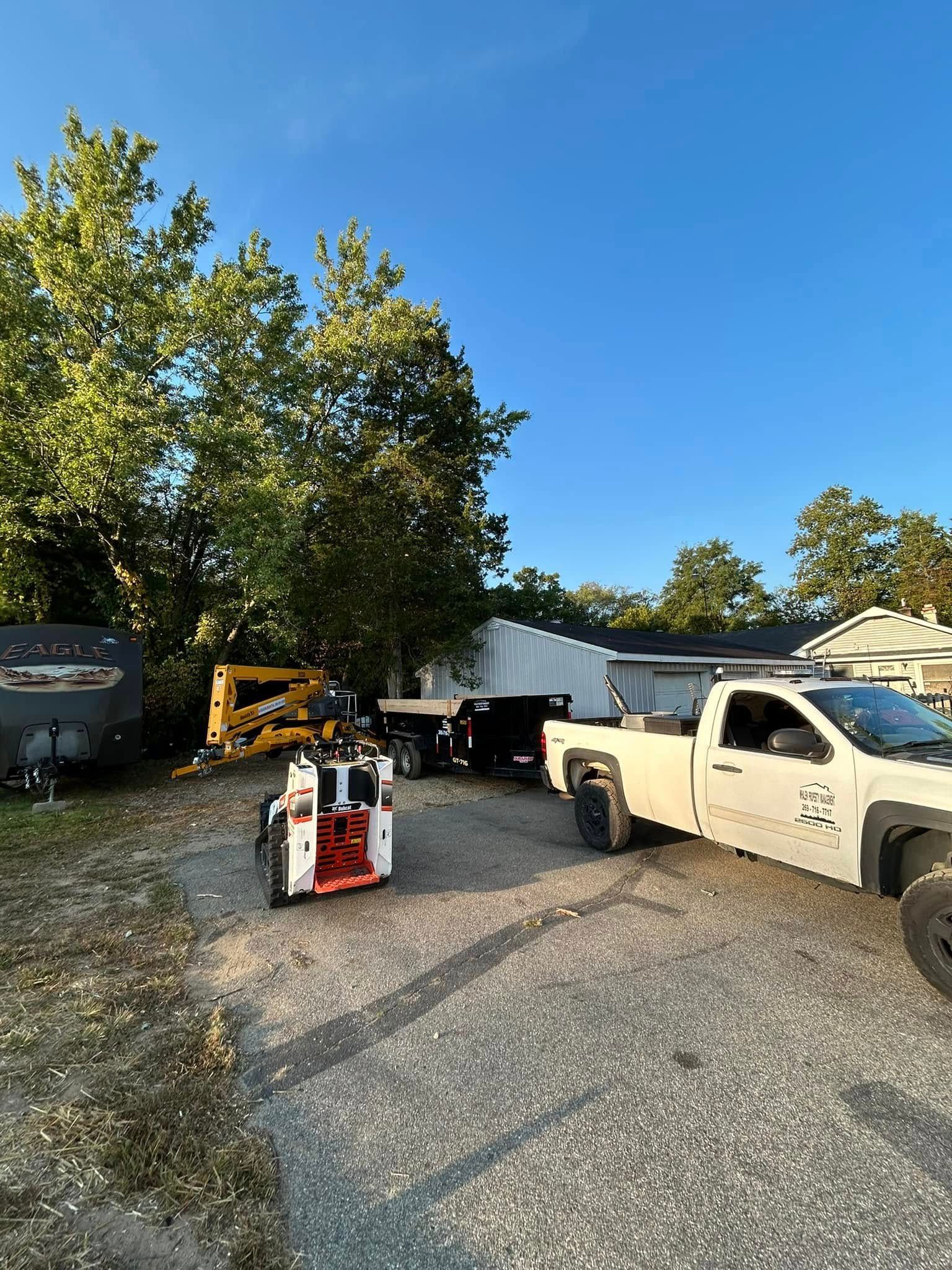 A white pickup truck, a skid steer, and a trailer parked on a gravel lot next to trees under a clear blue sky.