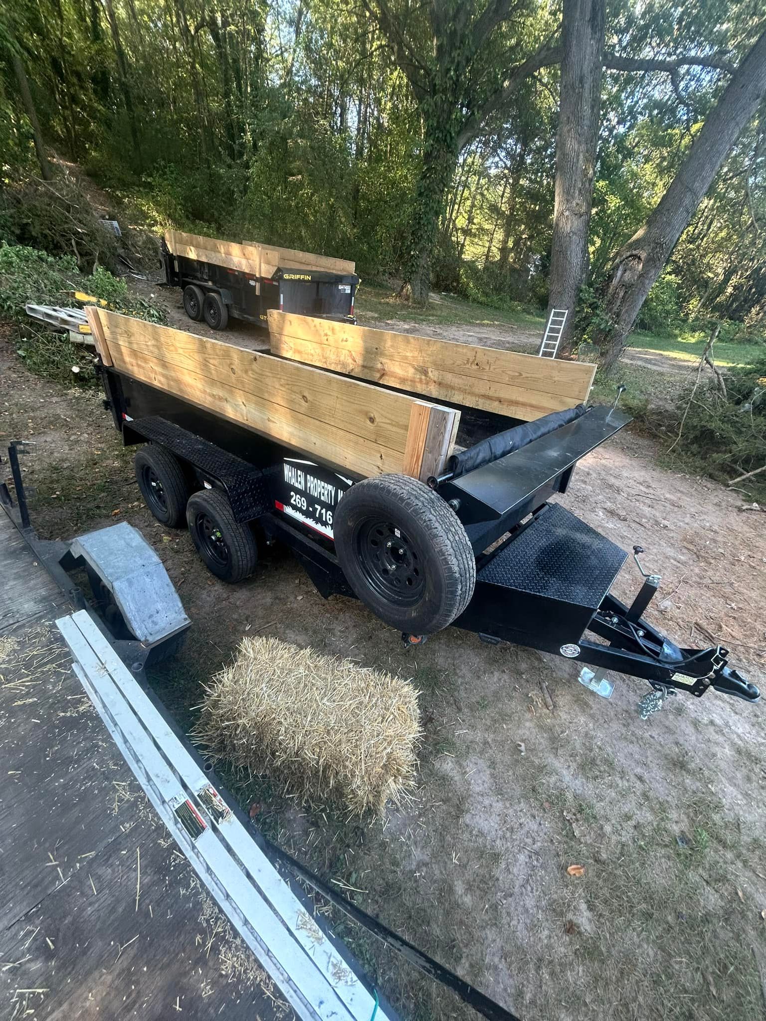 A black dump trailer with wooden side extensions parked on a dirt path in a wooded area.