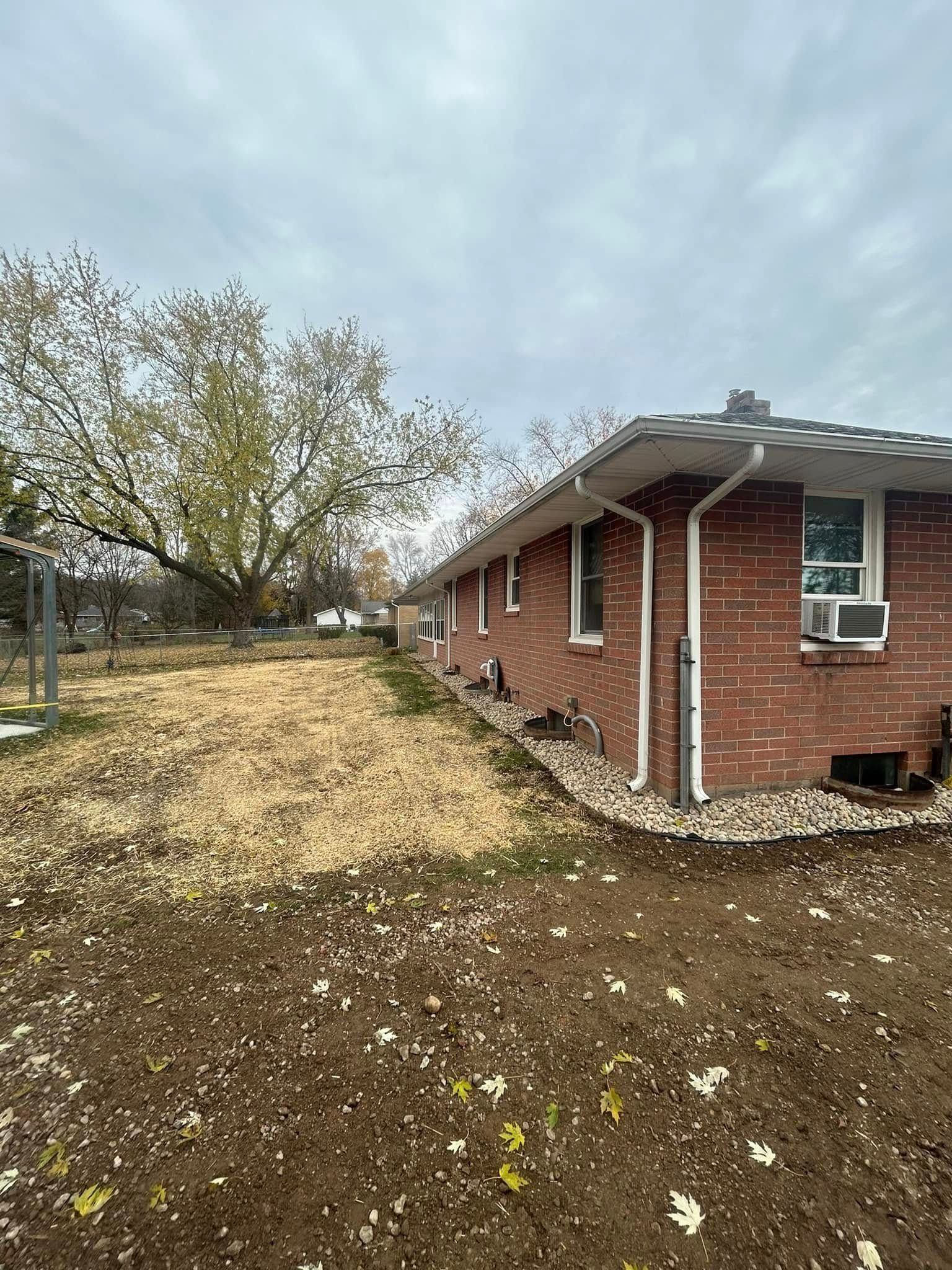 A side view of a red brick house with white trim under a cloudy sky, alongside a yard covered in fallen autumn leaves.