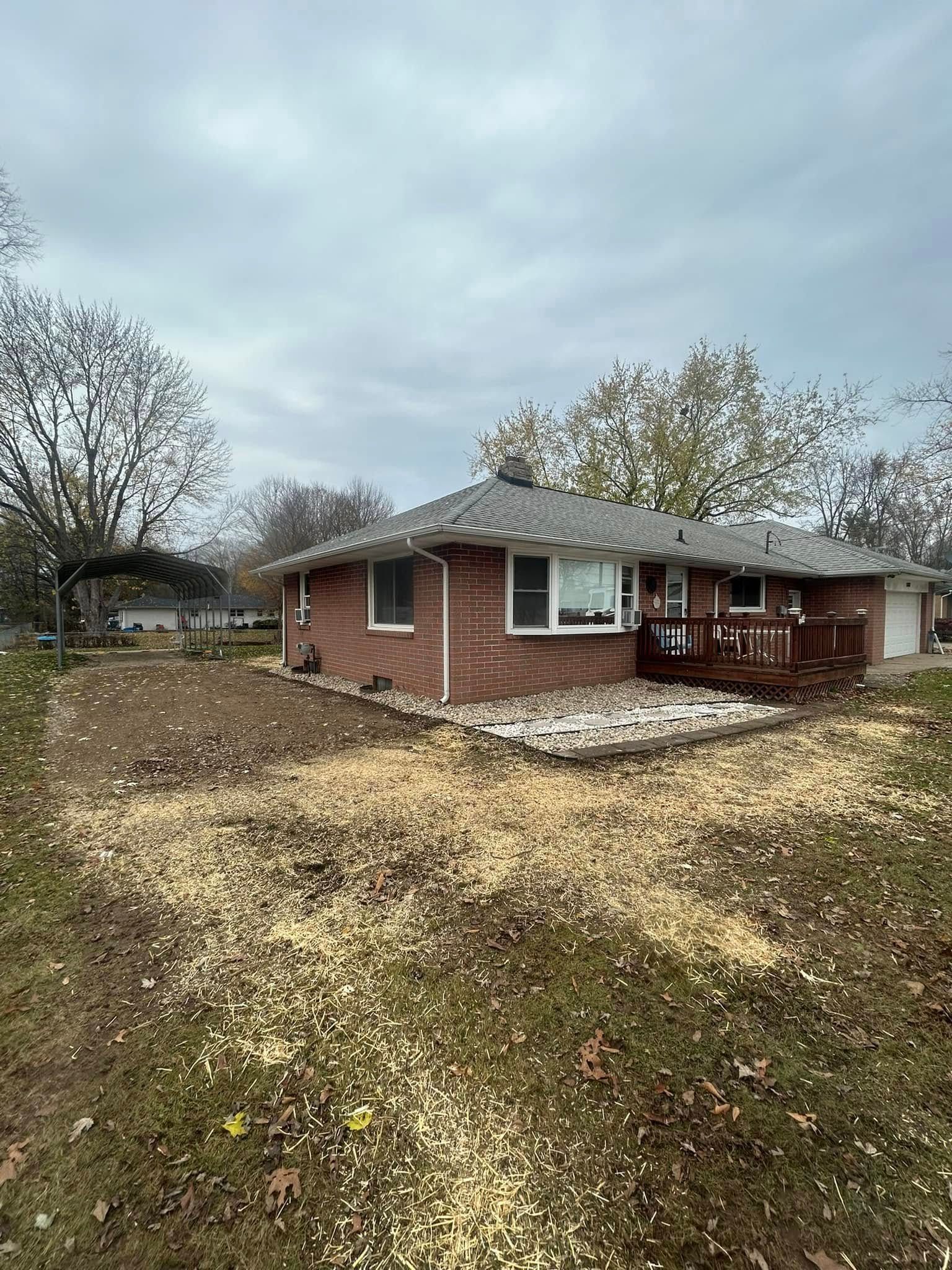 A single-story red brick house with a dark roof and wooden deck under a cloudy sky, surrounded by yard debris.