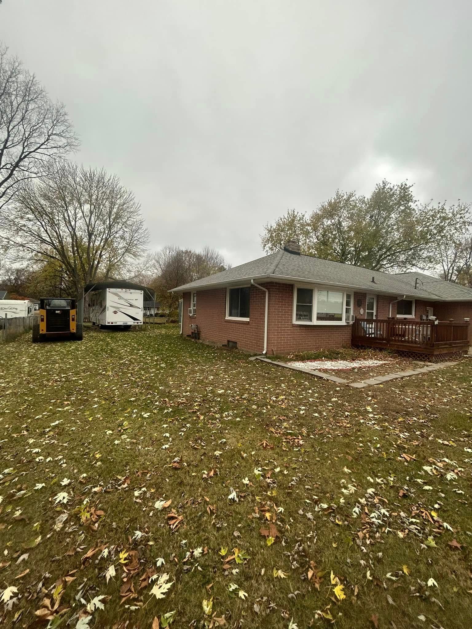 A brick house with a dark roof and a small deck sits on a grassy yard covered with autumn leaves under a cloudy sky.