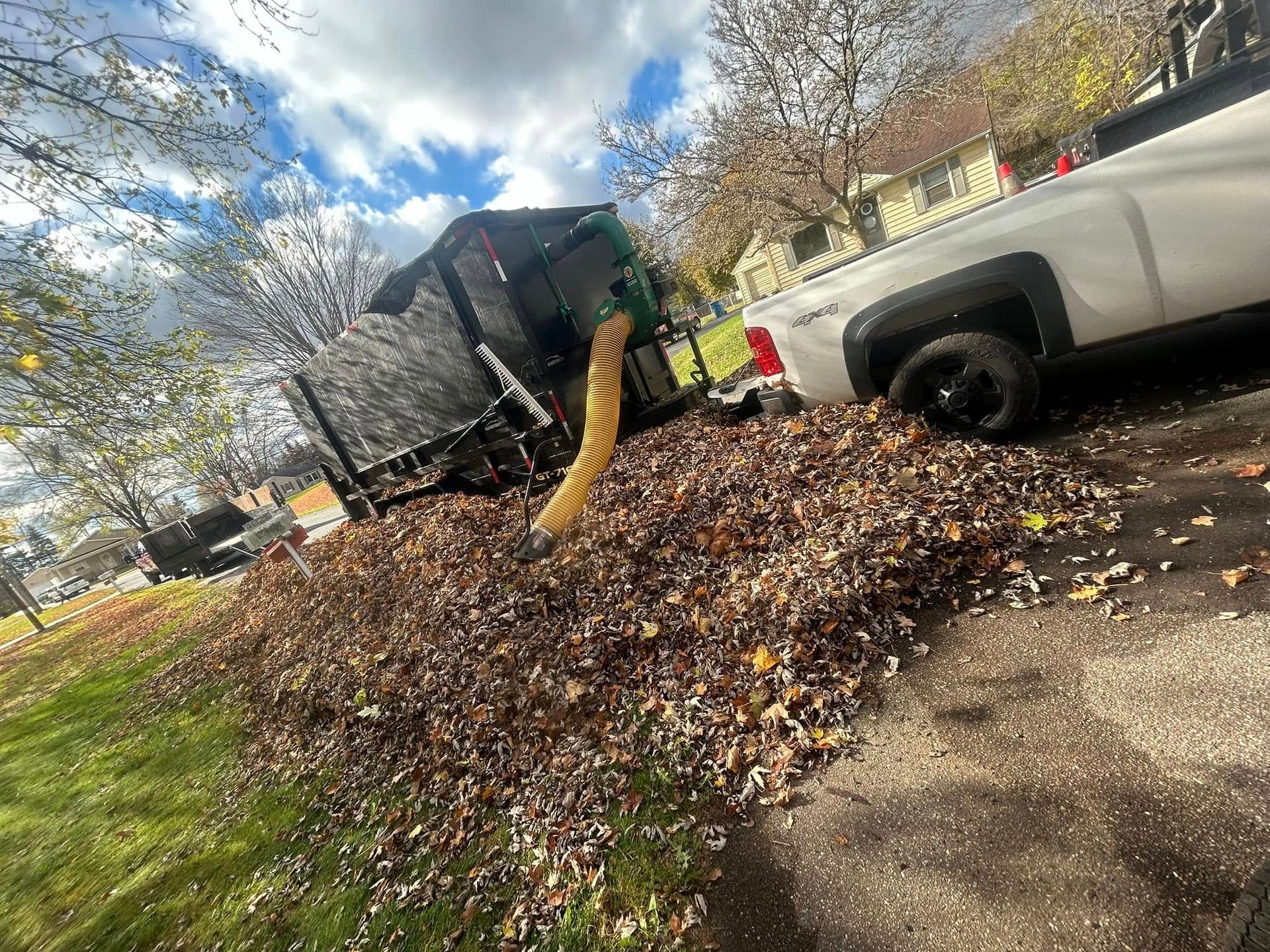 A white truck parked on a driveway with a large leaf vacuum machine collecting a massive pile of fallen autumn leaves.