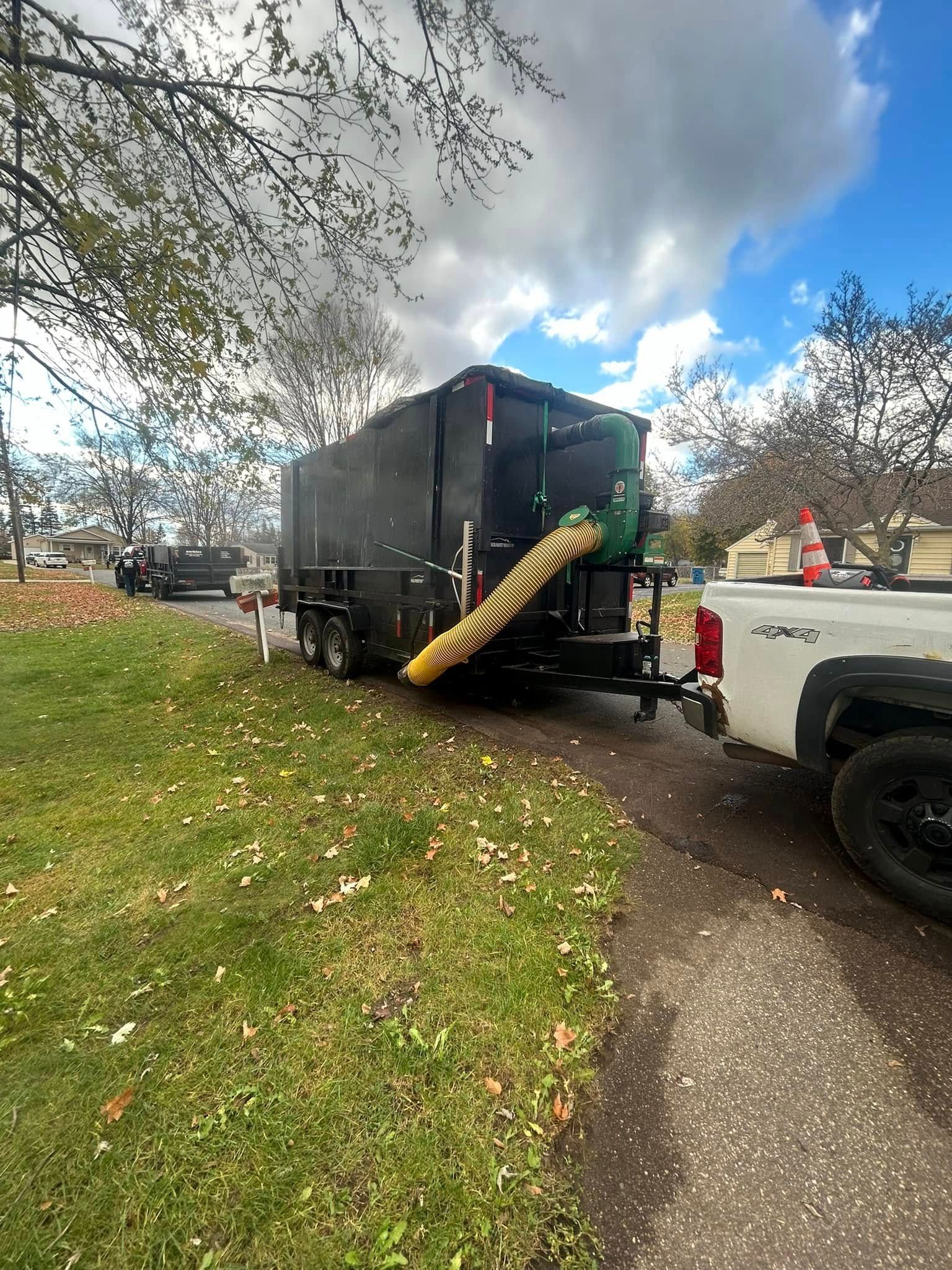 A white pickup truck tows a large, black leaf vacuum trailer along a grassy roadside on a partly cloudy day.