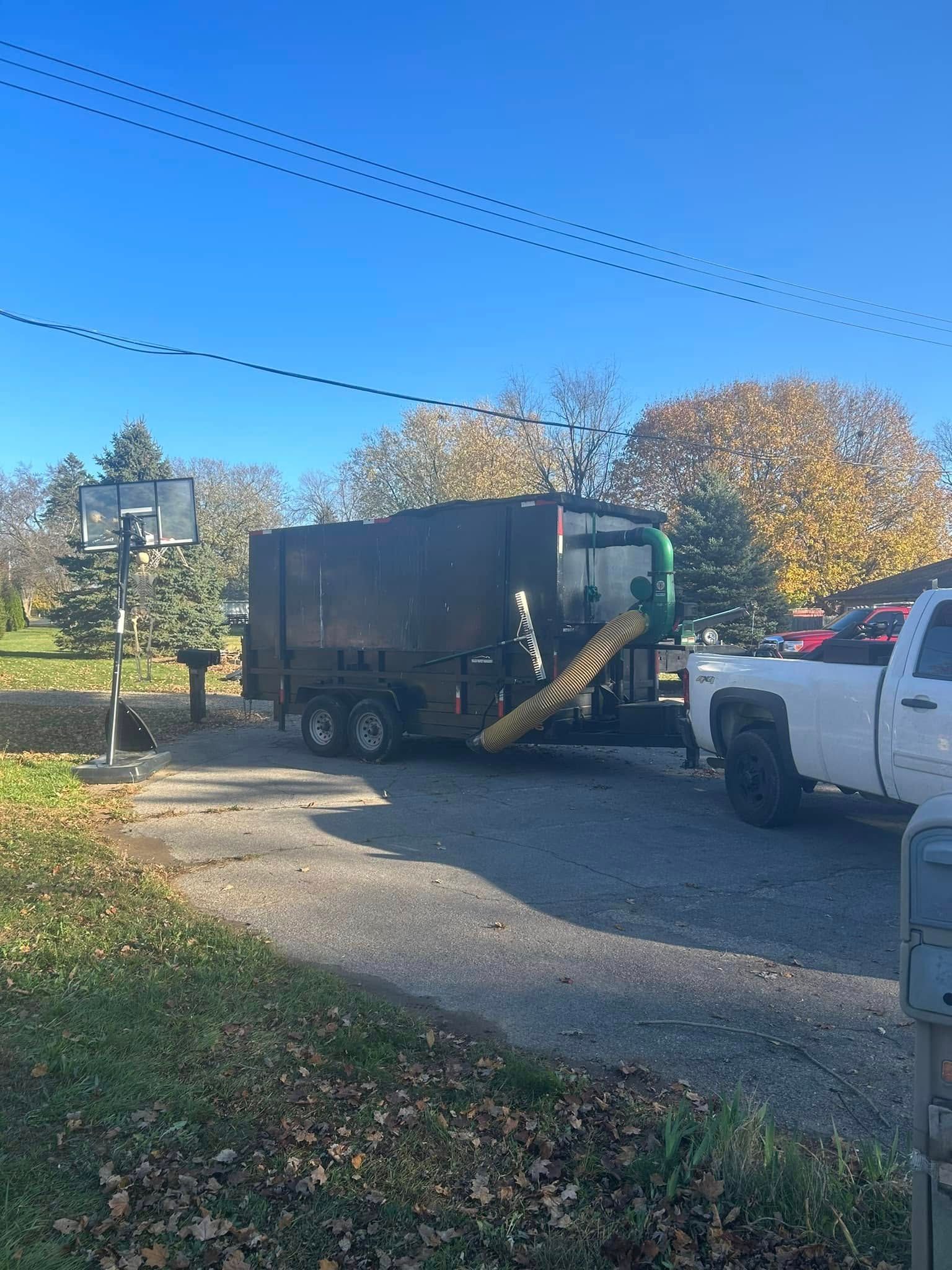 A white pickup truck pulls a black leaf vacuum trailer on a sunny, leaf-covered residential driveway.