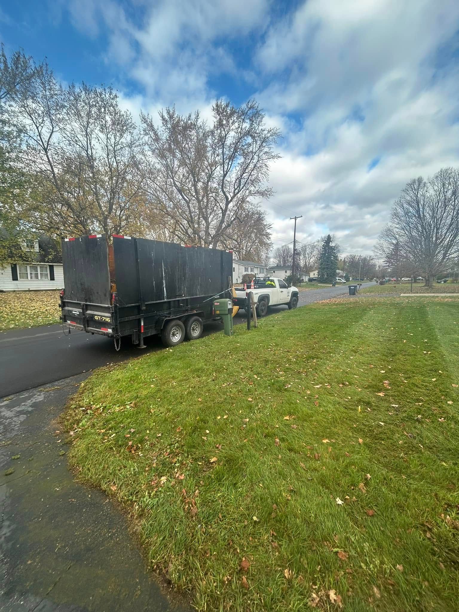 A white pickup truck pulling a large, black dump trailer parked on a suburban driveway near a grassy lawn.