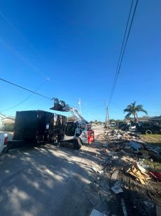 A skid-steer loader uses a grapple attachment to load debris from a residential street into a large dumpster.
