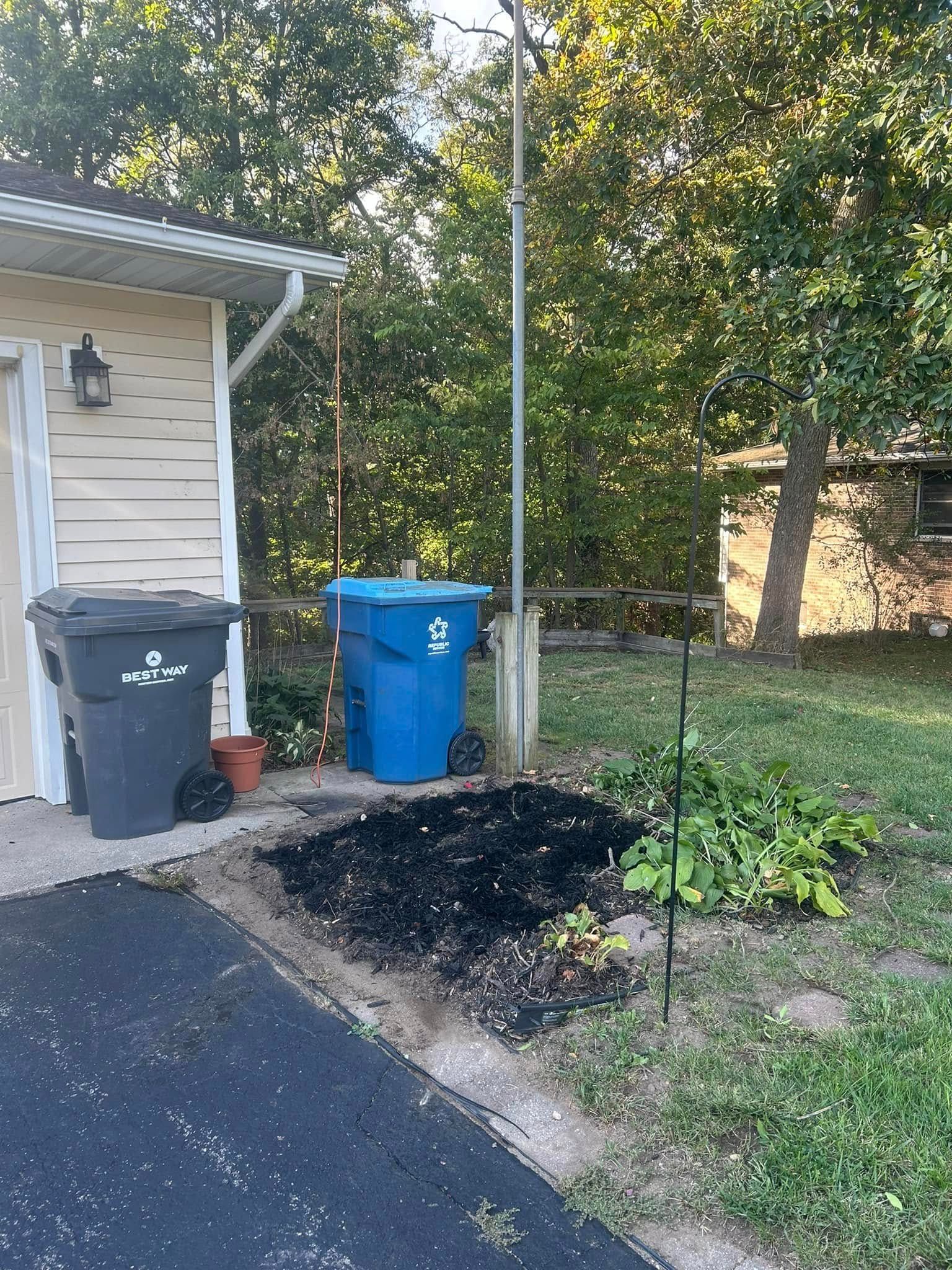 A light-colored house corner beside two garbage bins, a dark soil patch, and a green garden bed next to a metal pole.