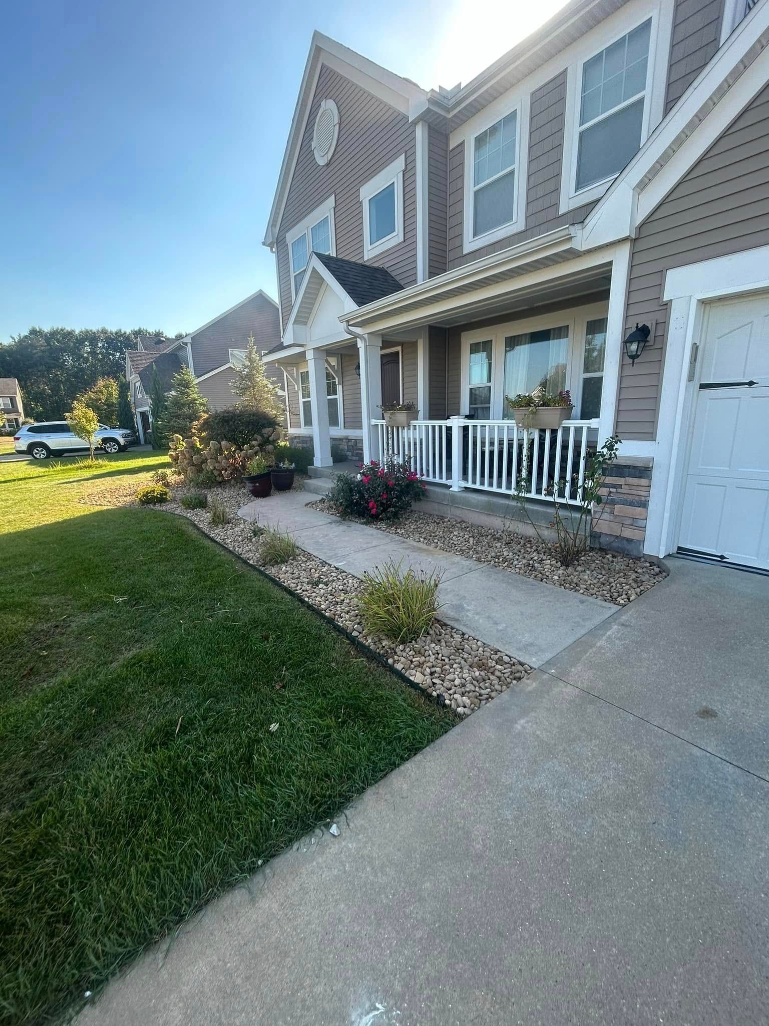A tan-sided suburban house with a white front porch, a stone-lined garden bed, and a concrete driveway on a sunny day.
