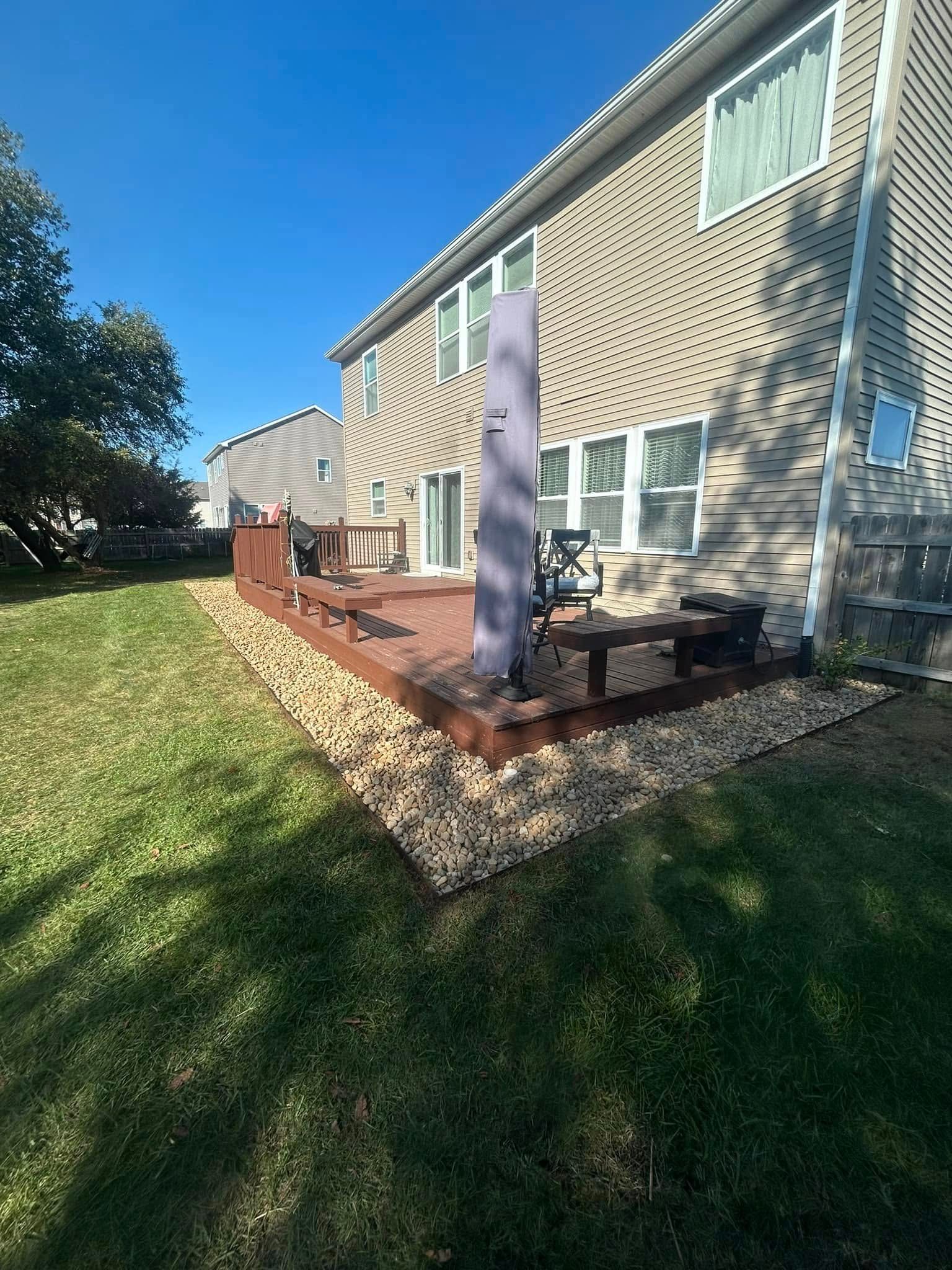 A wooden deck with patio furniture and a closed umbrella, bordered by decorative landscaping stones against a house wall.