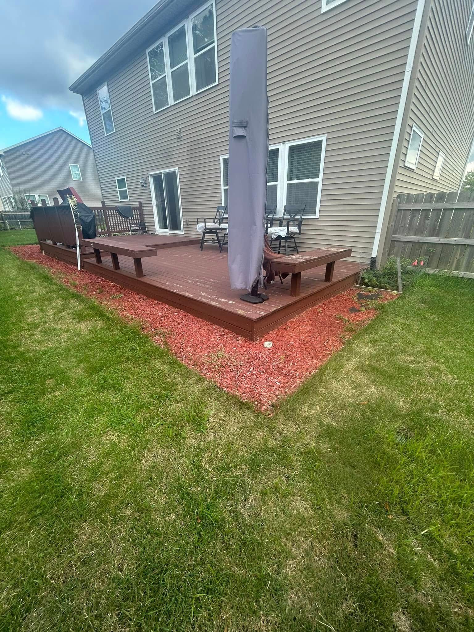 A backyard wooden deck with a covered patio umbrella, benches, and red mulch landscaping against a house with beige siding.