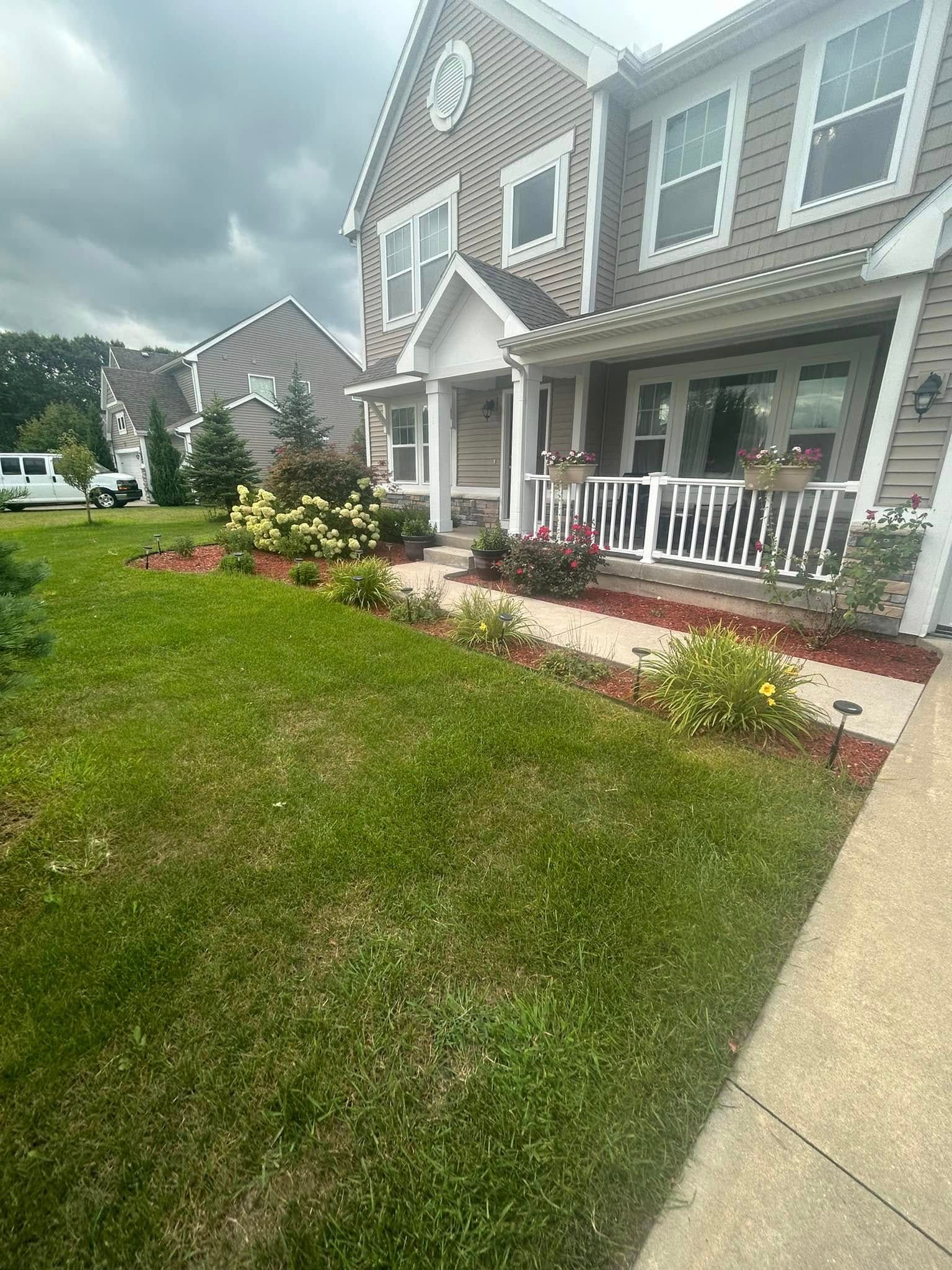 A multi-story beige house with a front porch, green lawn, and landscaped garden beds under a cloudy sky.