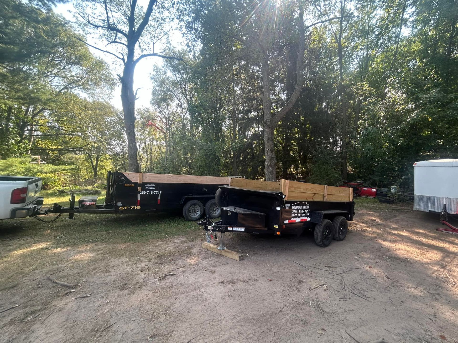 Two black dump trailers with wooden side extensions parked on a dirt lot in front of trees.
