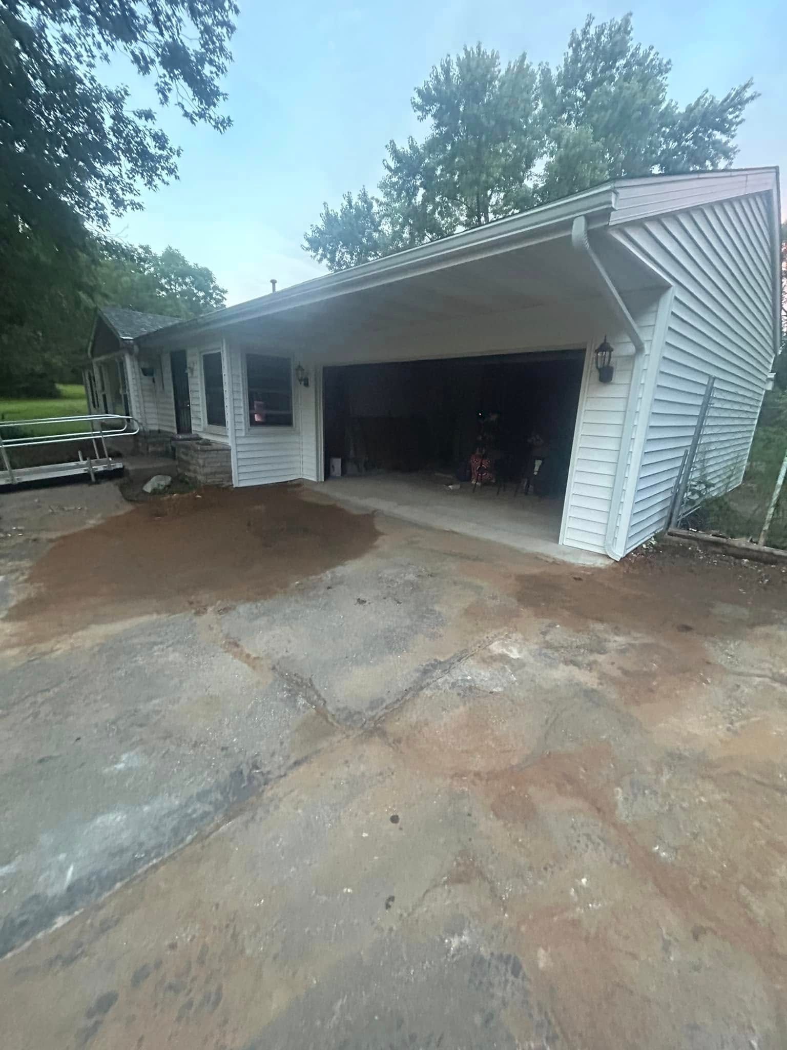 A white house with a carport and a patch of dirt on the concrete driveway, surrounded by trees under an overcast sky.