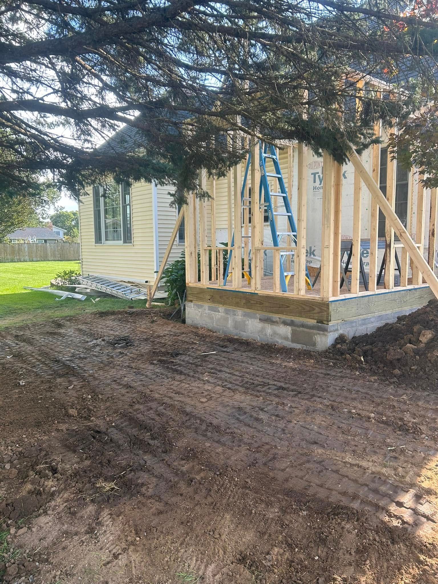 Wooden wall framing for a house addition sits on a concrete foundation next to an existing yellow house.