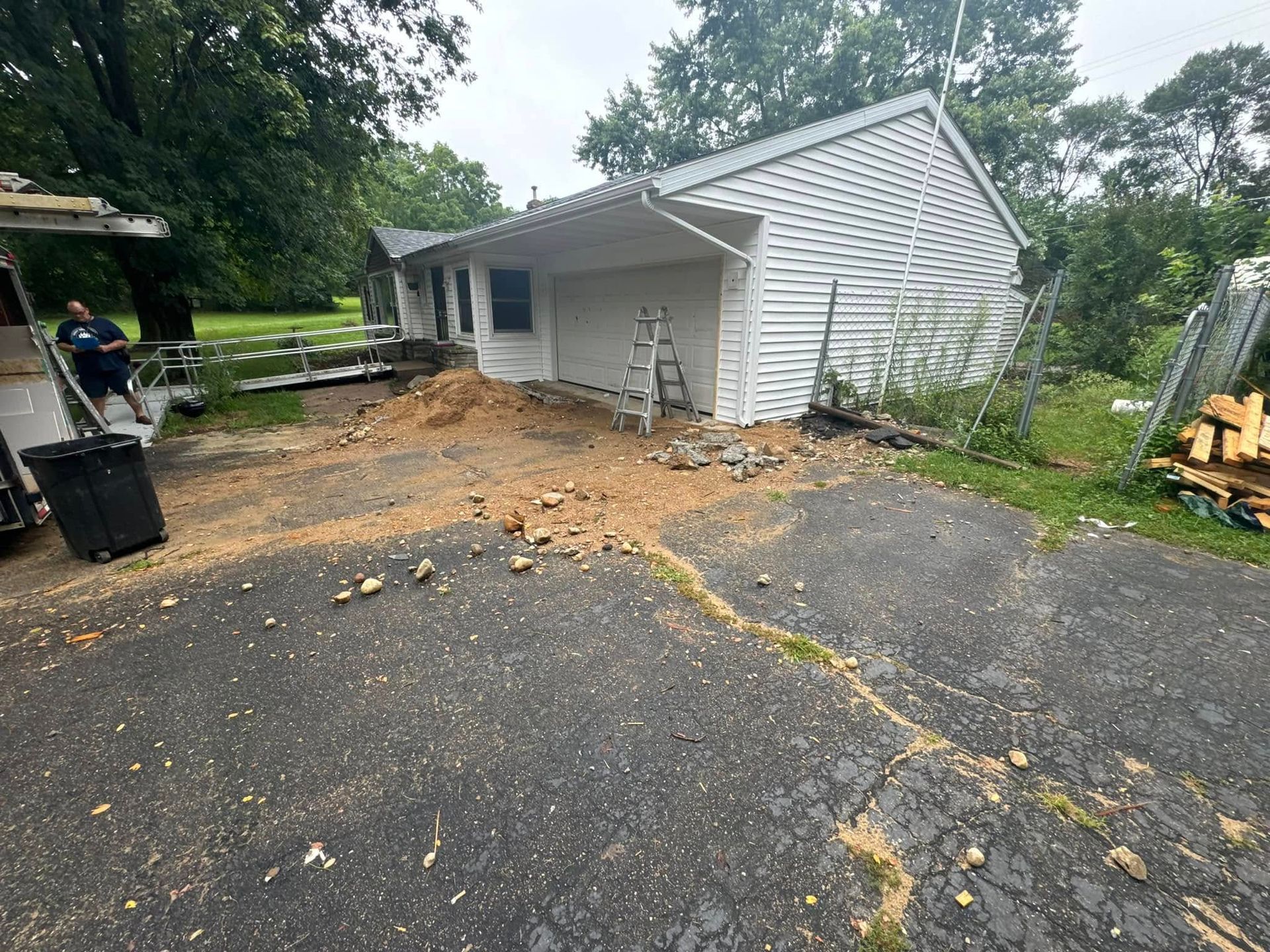 A person stands near a white house with a garage, with a pile of dirt and debris covering the driveway in front of it.