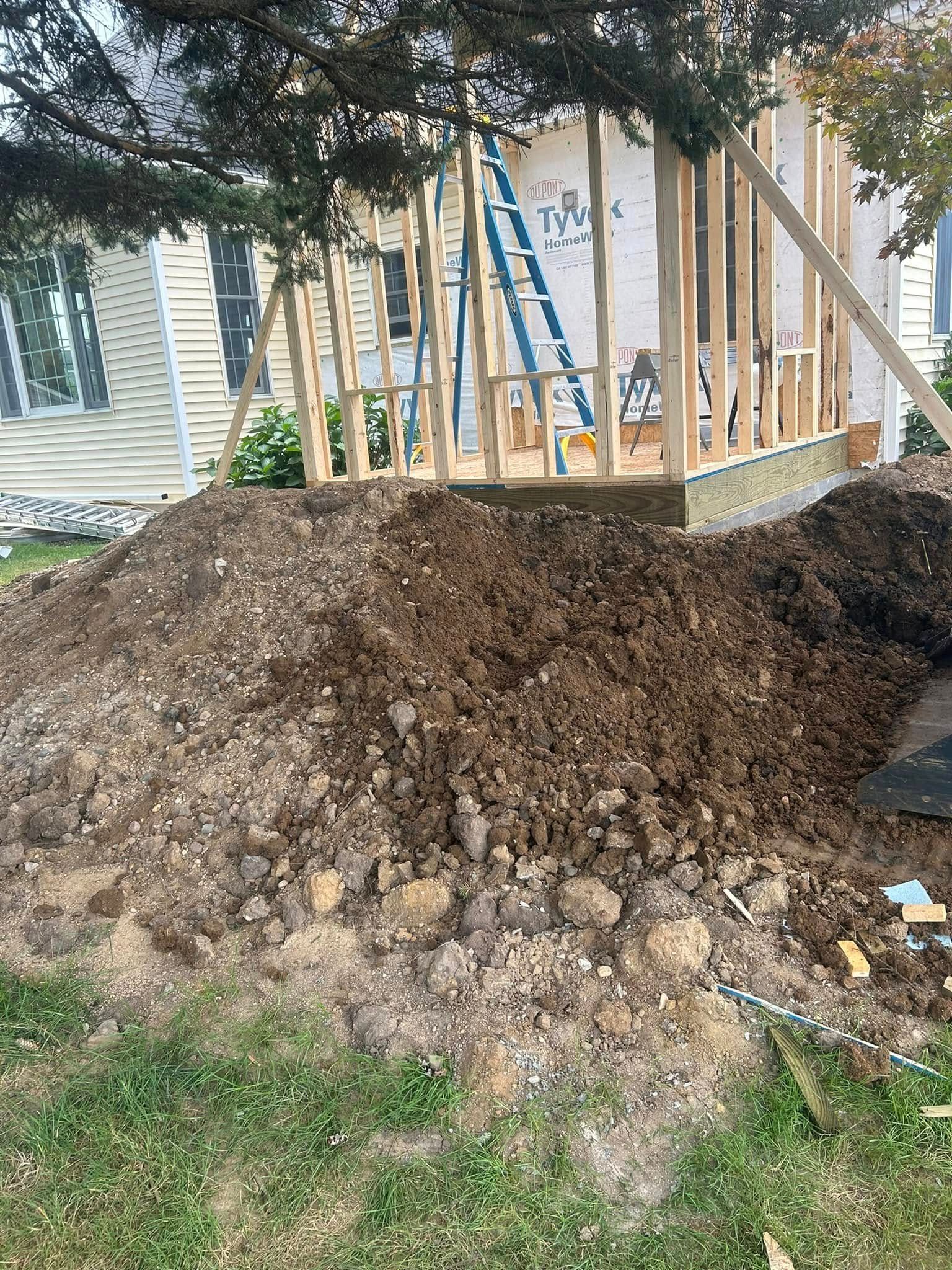A pile of excavated dirt sits in front of a wooden house framing structure with a blue ladder leaning against it.