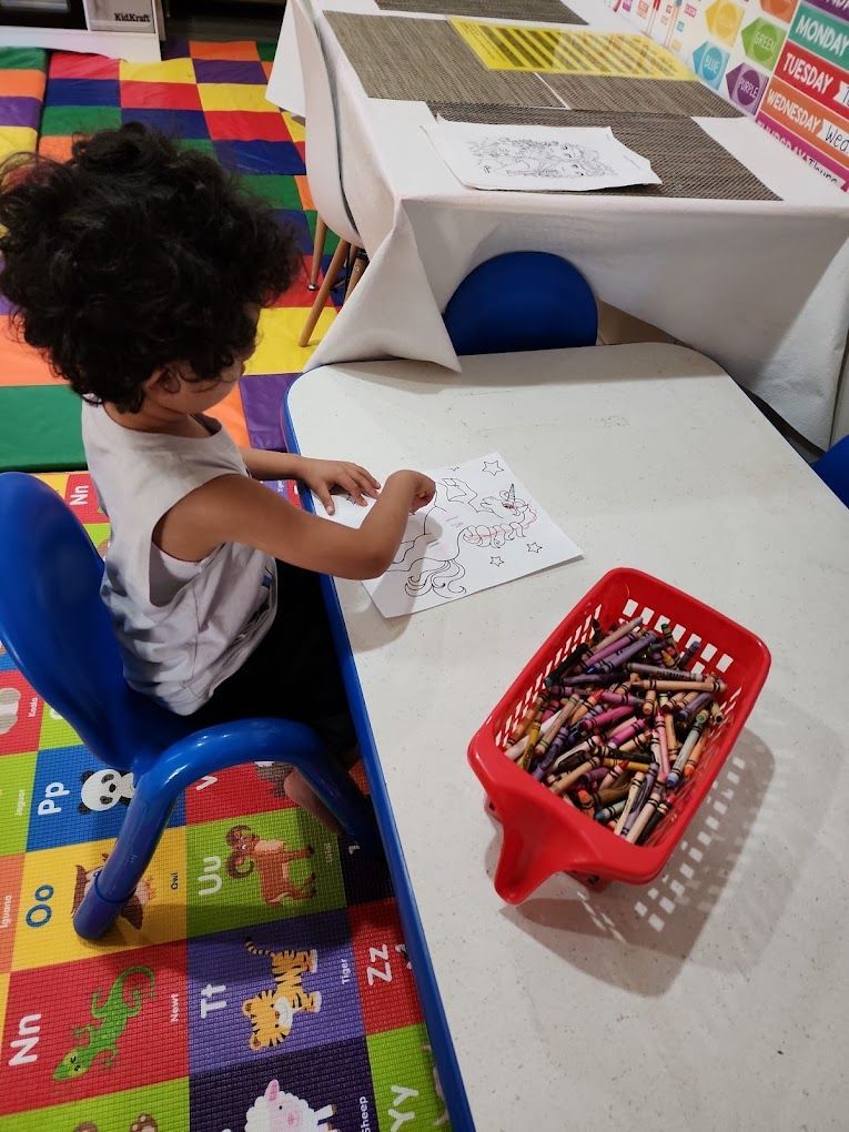 Child seated, drawing at a white table with crayons in a red basket. Colorful floor.