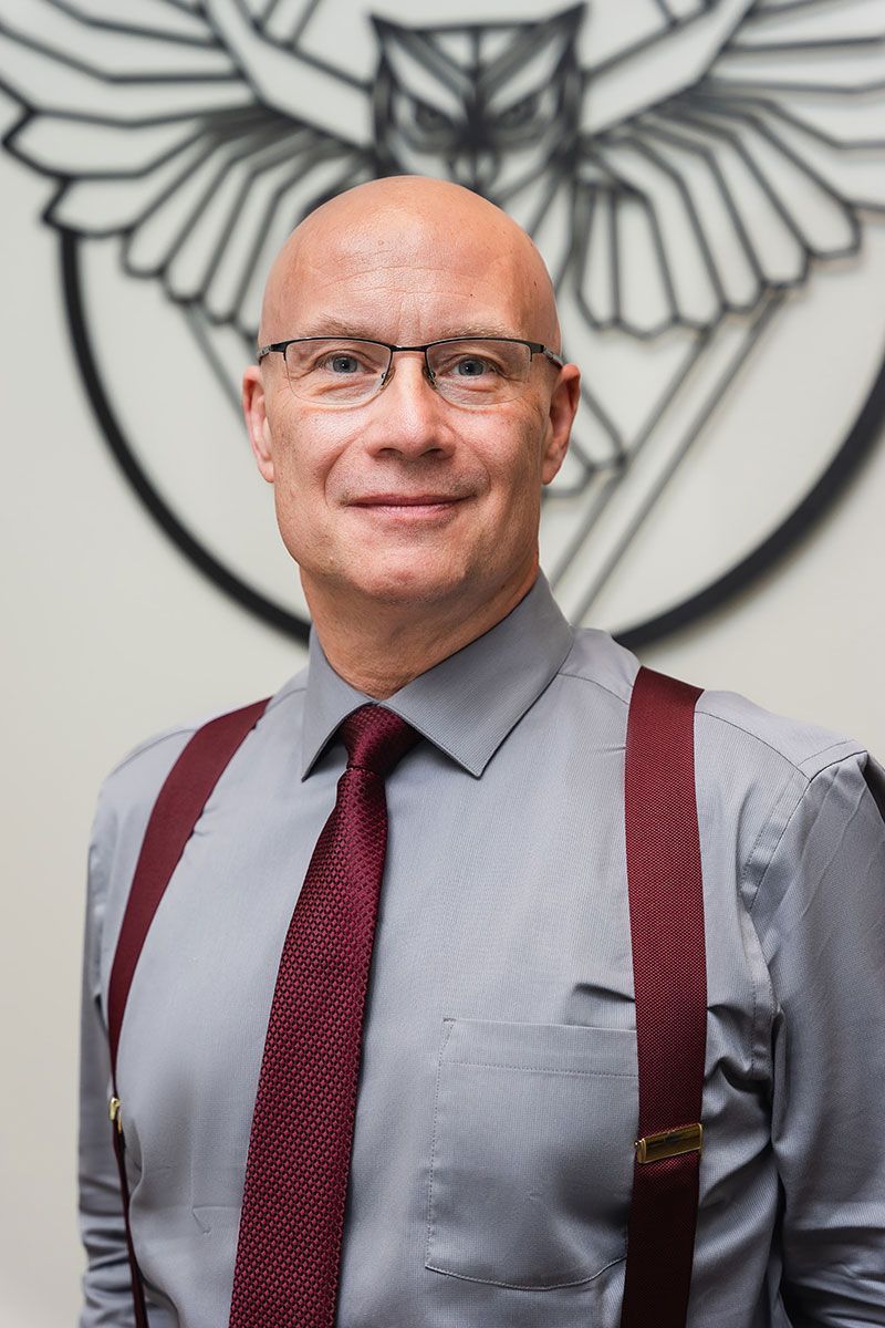 Bald man in glasses wearing gray shirt, red tie, and suspenders, standing before a winged emblem backdrop