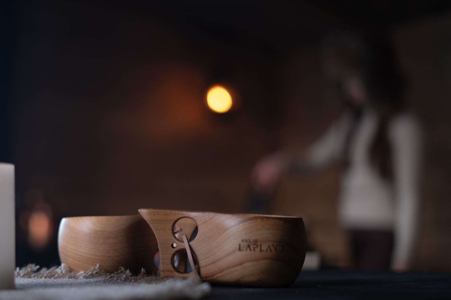 Kuksa wooden mugs in front of woman cooking in the back ground in candle light.