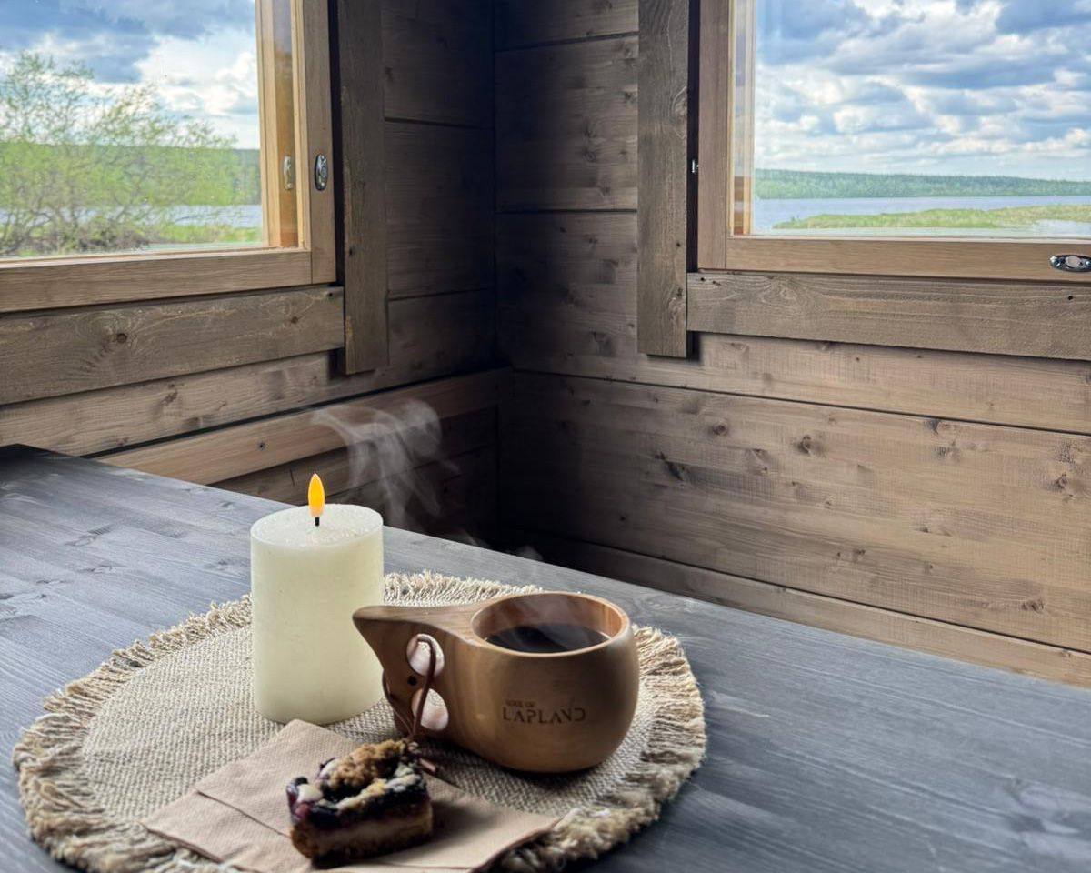 Kuksa mug with coffee on a log cabin table with windows opening to lush green river views