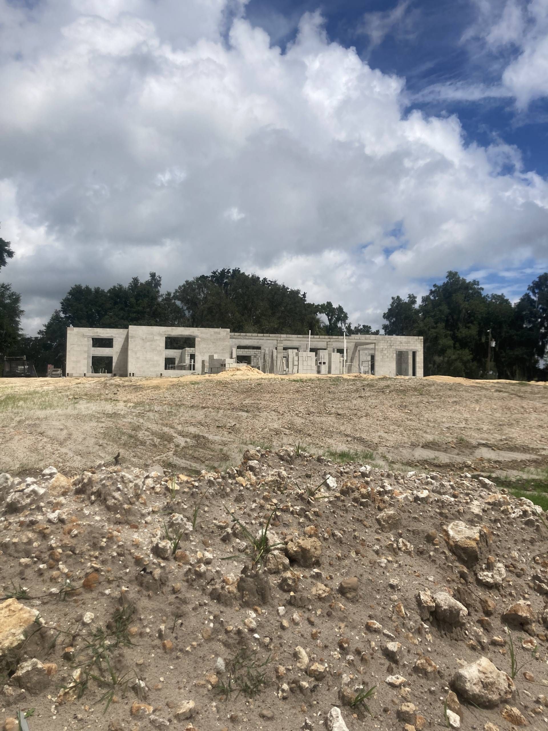 A low concrete building under construction behind a rocky dirt lot, with cloudy sky and trees in the background