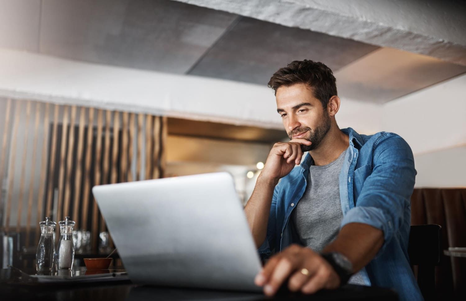 Homem em frente ao laptop, em um café, olhando pensativamente para a tela, com a mão no queixo.
