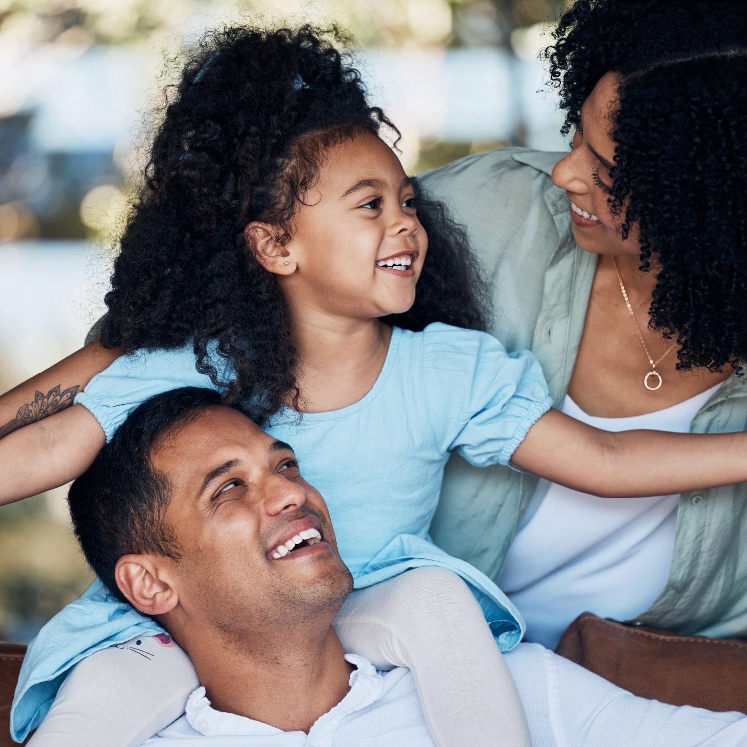 Família sorrindo, filha nos ombros do pai, mãe por perto. Ao ar livre, expressões felizes, cores claras.