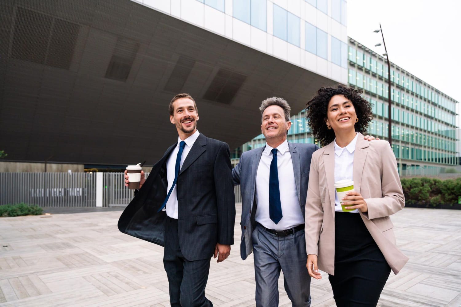 Três empresários caminhando e sorrindo do lado de fora de um prédio comercial moderno, segurando xícaras de café.