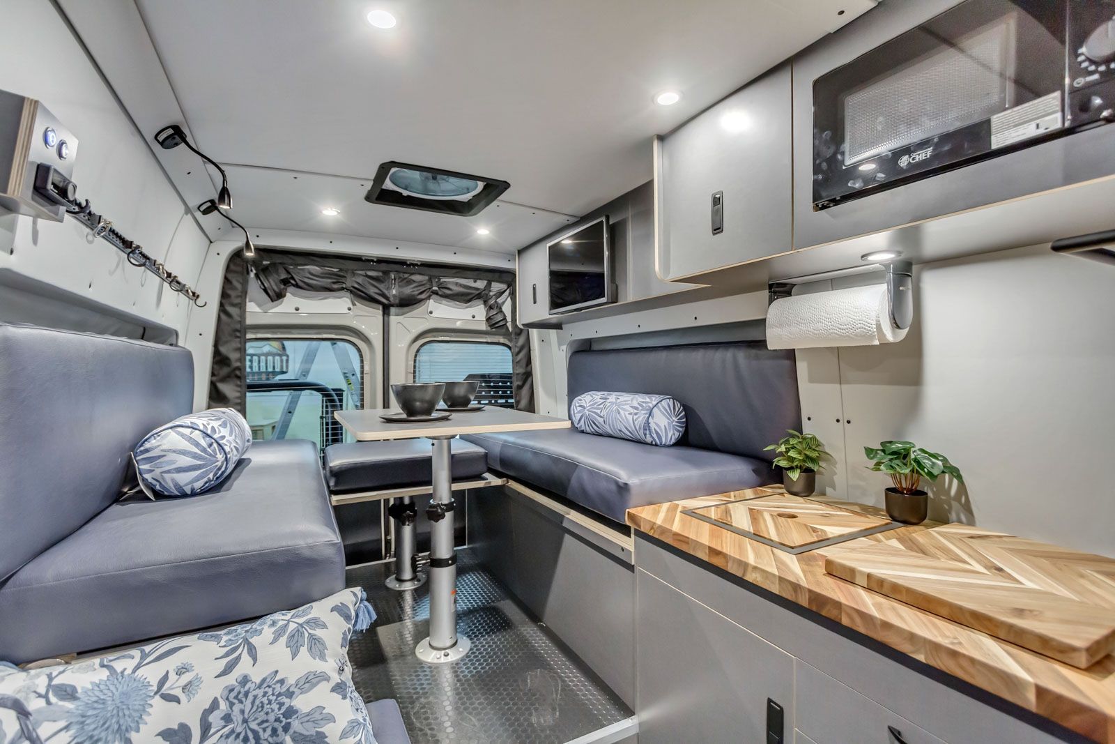 Modern camper van interior with gray seating, wood countertop, and compact kitchen/storage cabinets