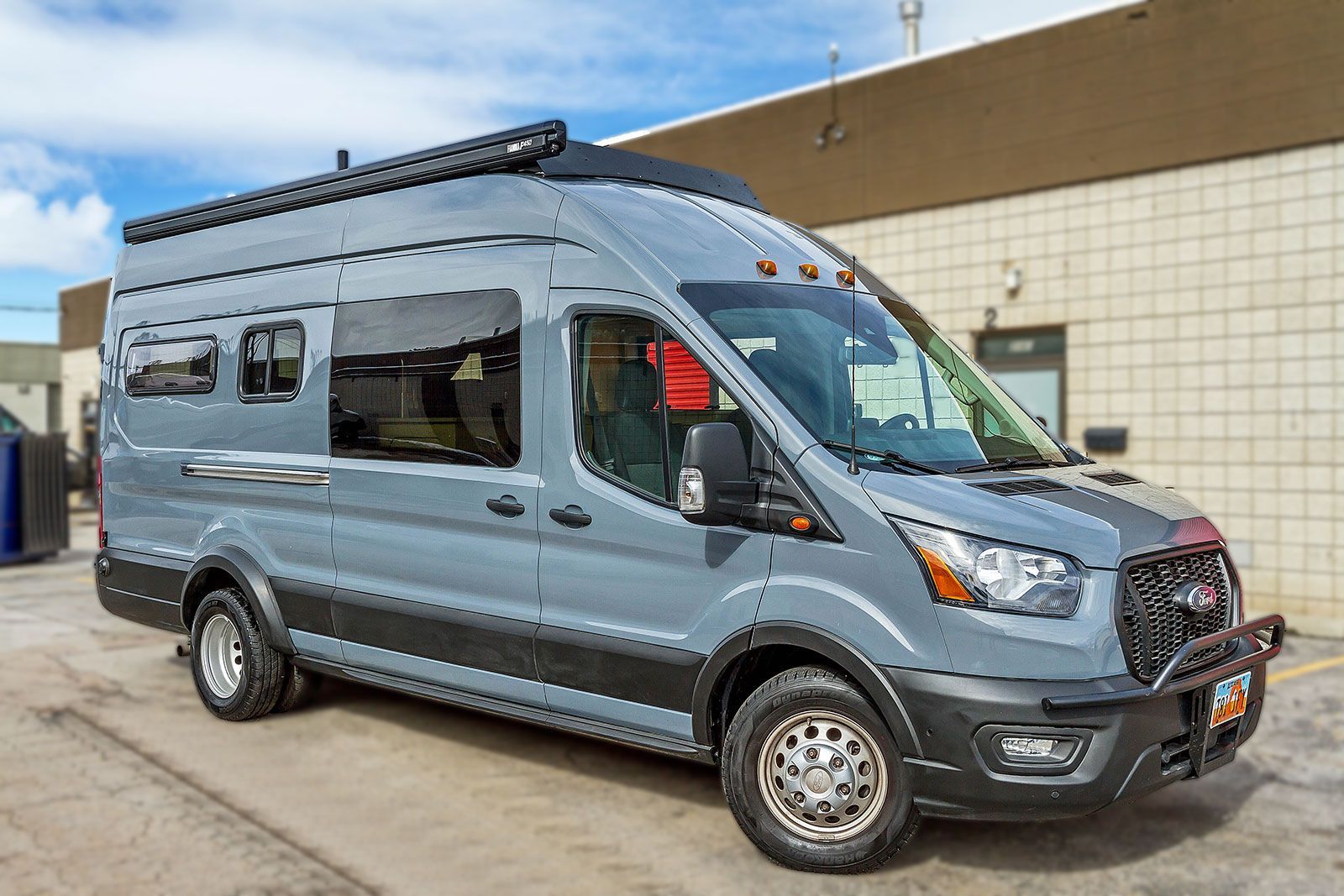 Blue-gray camper van parked outside a building under a clear sky.