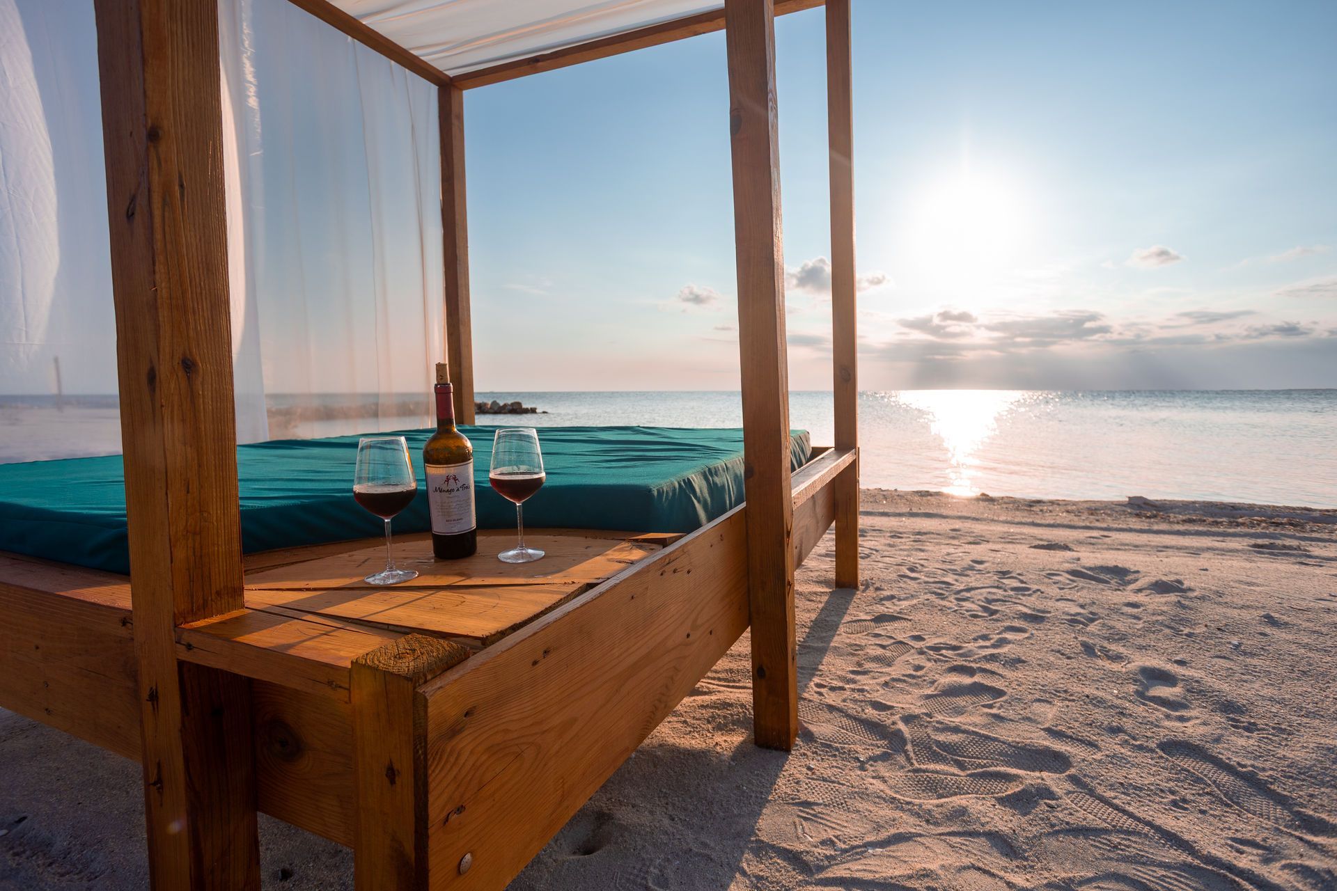 Beach cabana with a small table, bottles, and turquoise cushions at sunset by the sea