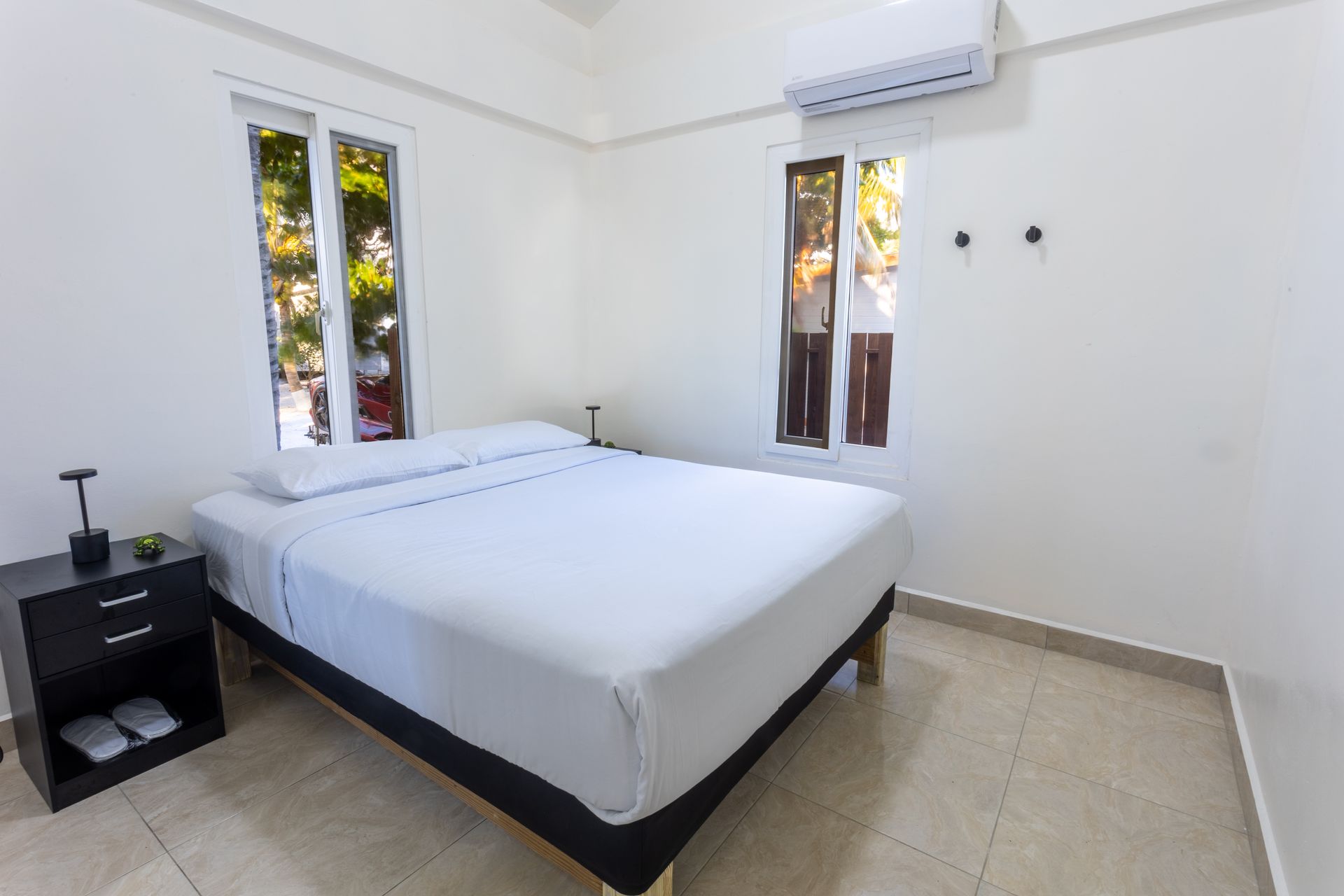 Minimal bedroom with a white bed, nightstand, windows, and wall-mounted air conditioner.