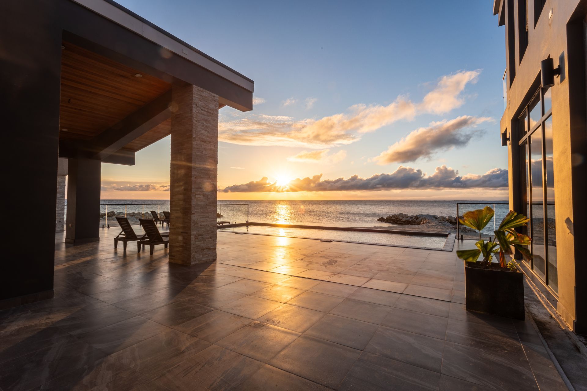 Sunset over a seaside patio with reflective wet tiles and ocean waves beyond.