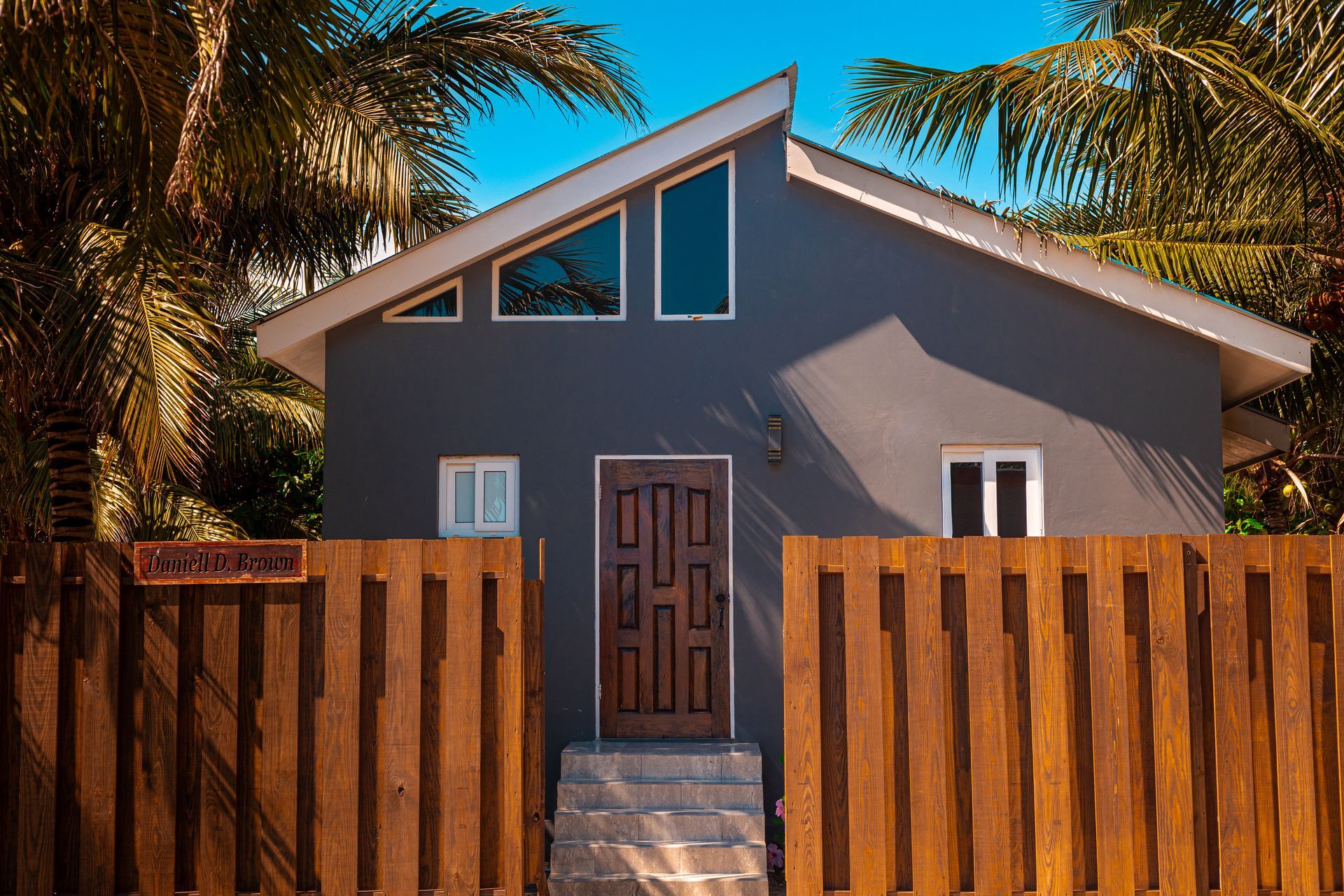Gray house behind a wooden fence with a centered front door and palm trees at sunset