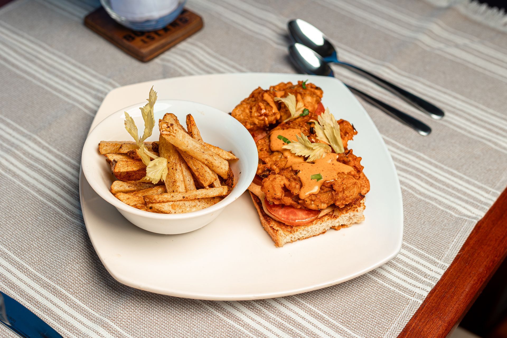 Plated sandwich with fries on a table, garnished with herbs, beside a spoon and fork