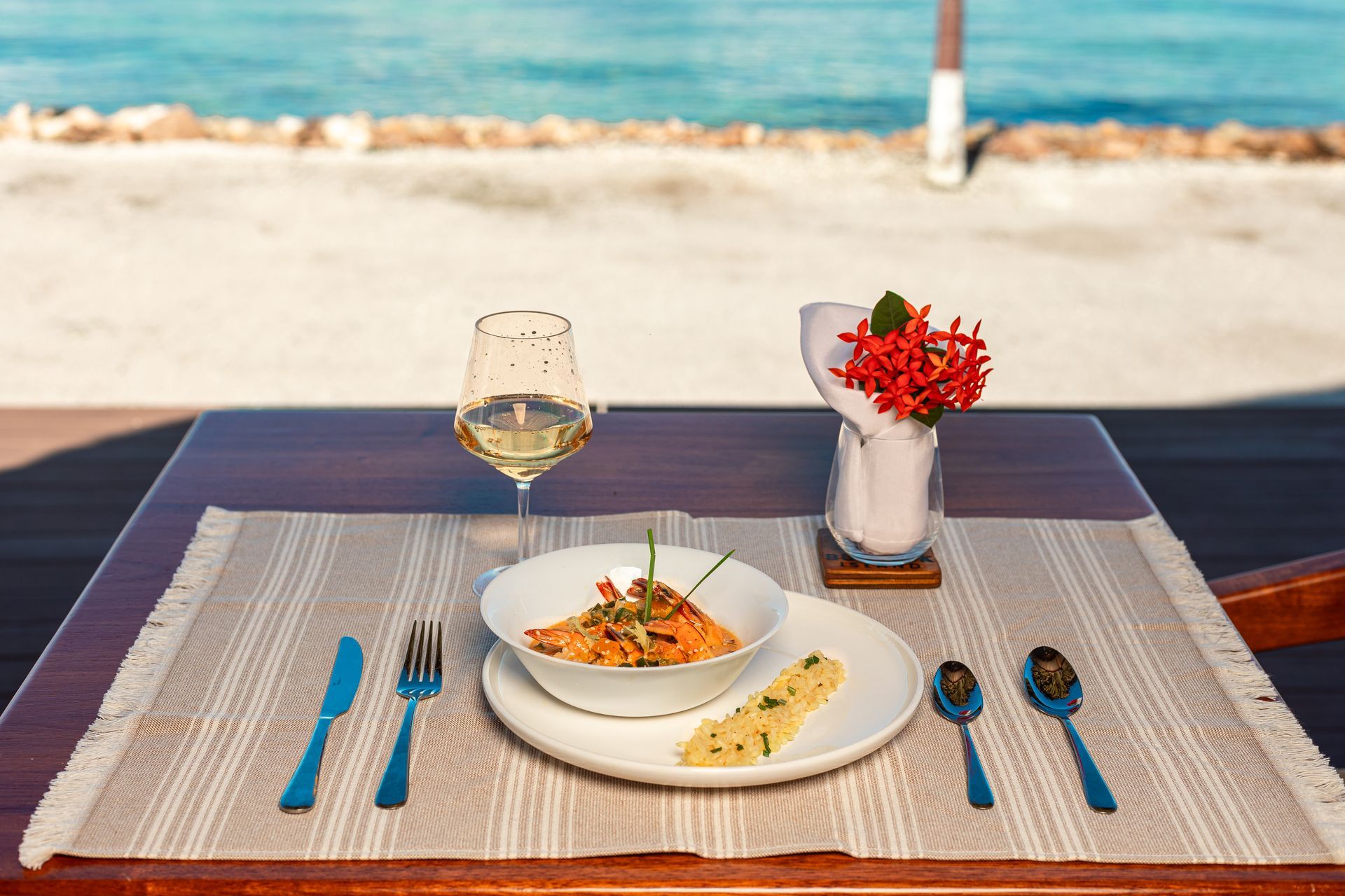 Beachfront table set with pasta, wine, flowers, and blue cutlery on a woven placemat
