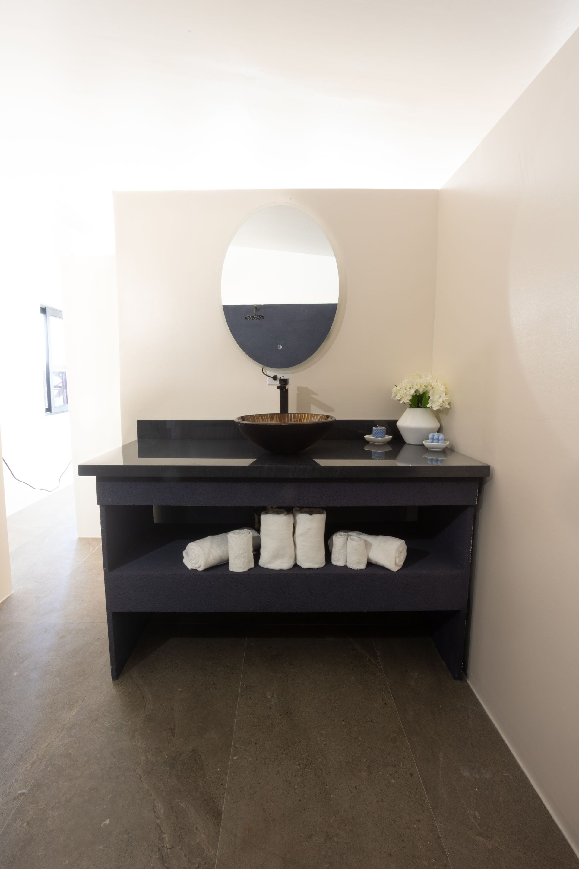 Modern bathroom vanity with oval mirror, dark counter, rolled towels, and flowers on a cream wall