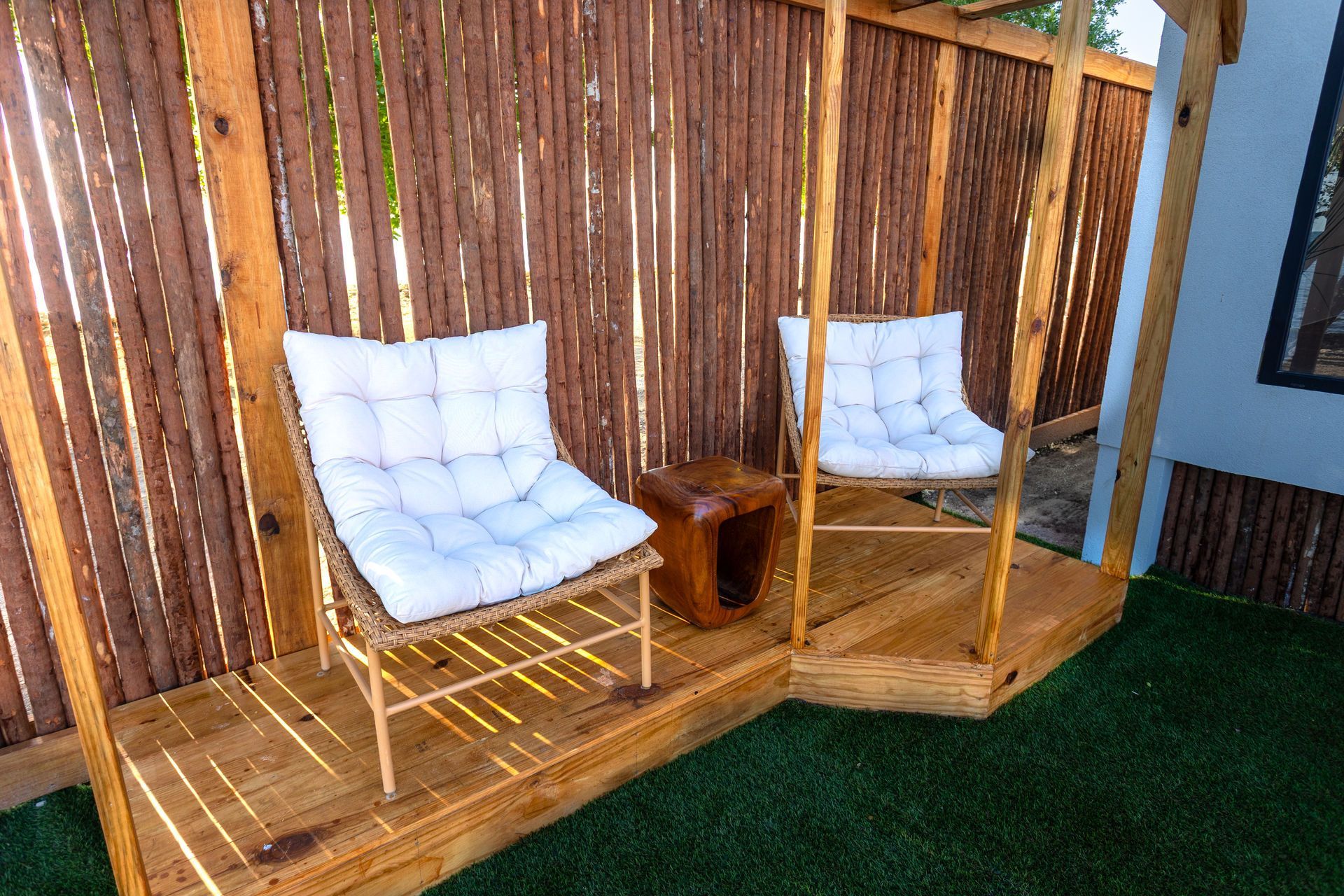 Two white cushioned chairs with a small wooden table on a deck beside a bamboo fence and grass.