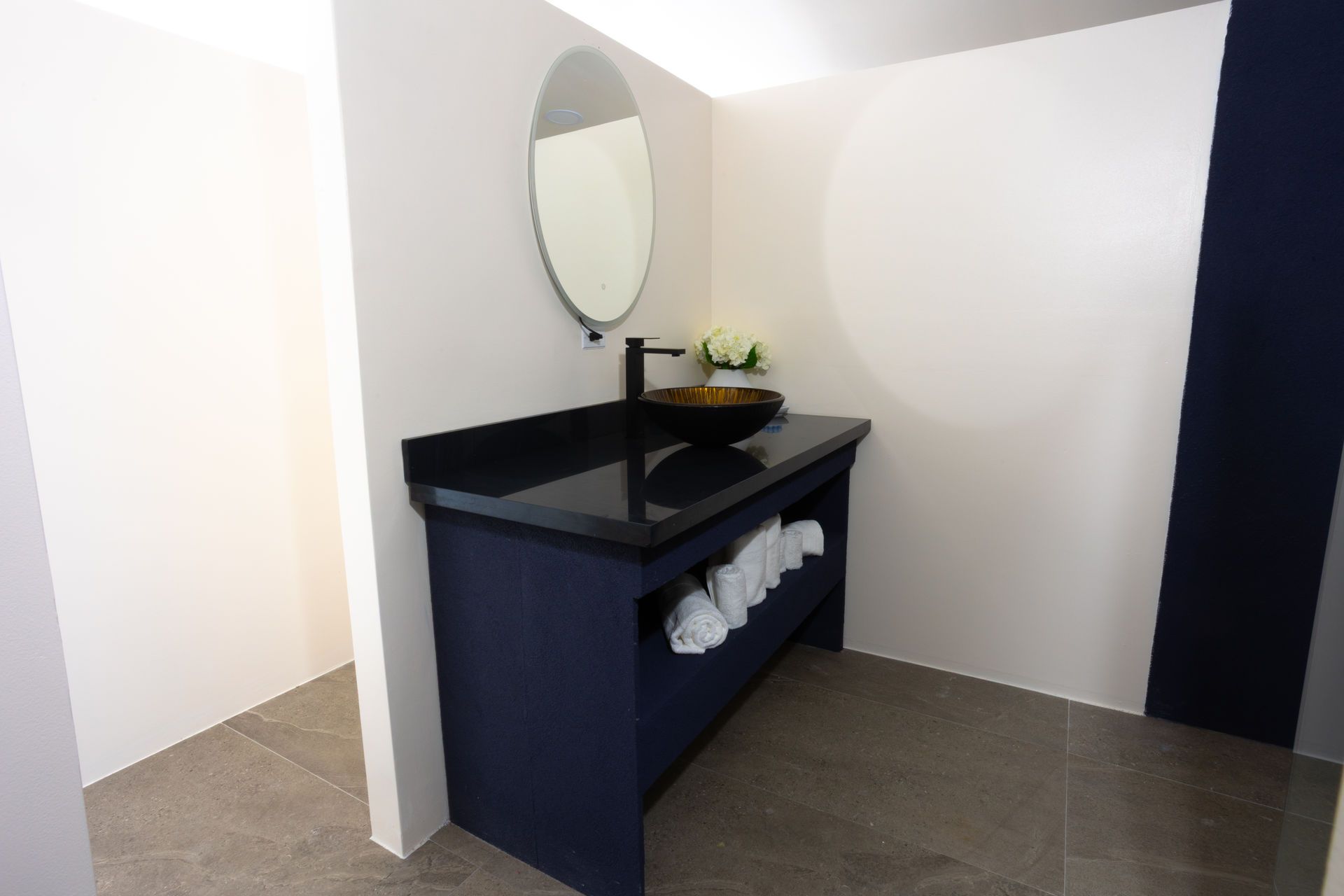 Modern bathroom vanity with round mirror, dark cabinet, countertop, and white towels on a wall shelf