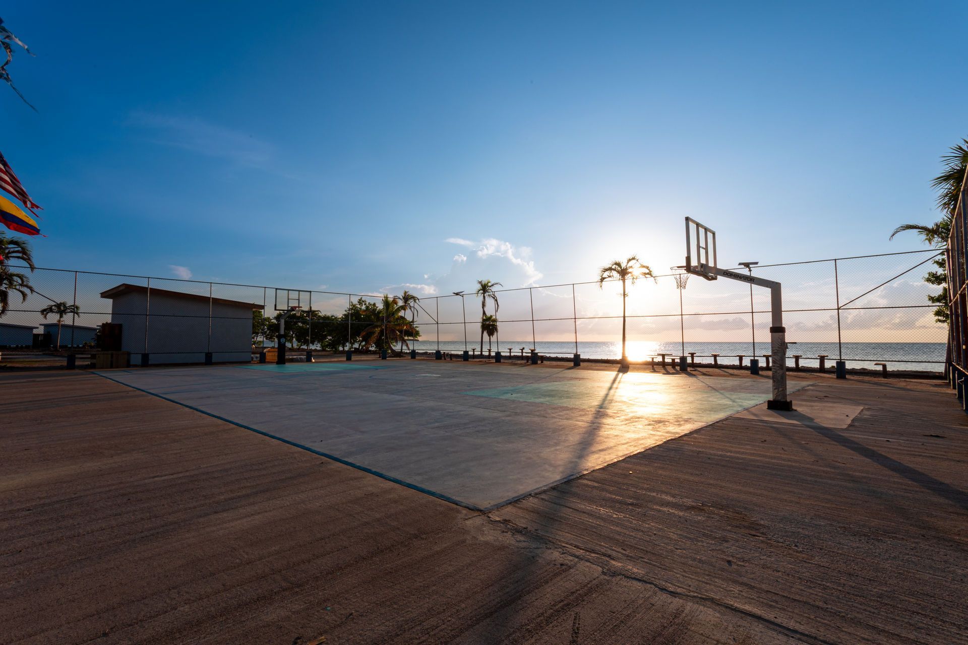 Sunset over an empty outdoor court with a basketball hoop, palm trees, and ocean in the background