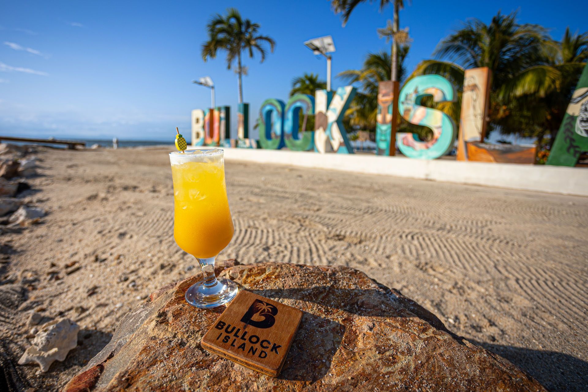 Yellow cocktail on a beach in front of the colorful “BULLDOG ISLA” sign and palm trees